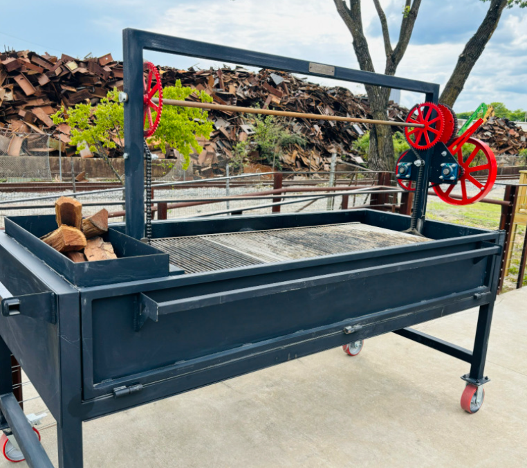 Large black metal firewood processor with red gears and a green wheel, placed outdoors on concrete, with a stack of cut firewood and piles of scrap metal in the background.