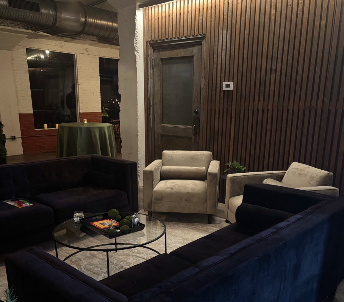 A cozy living room with dark and light upholstered armchairs and a glass coffee table in the center, decorated with small plants and candles. There is a textured wooden wall, a rustic door, and a high table with candles on a green tablecloth in the b