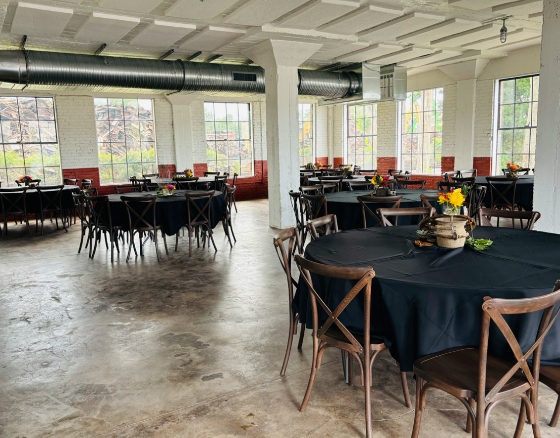 Indoor event space with round tables covered in black tablecloths, decorated with small flower arrangements, and wooden chairs. Large windows let in natural light, and the space has an industrial style with exposed ductwork and white brick walls.