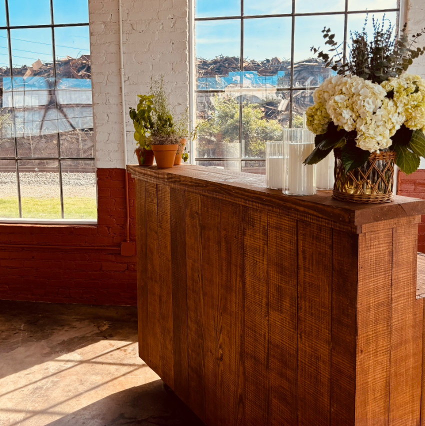 Interior space with large industrial-style windows, a white brick wall, and a wooden sideboard decorated with potted plants and white flowers.