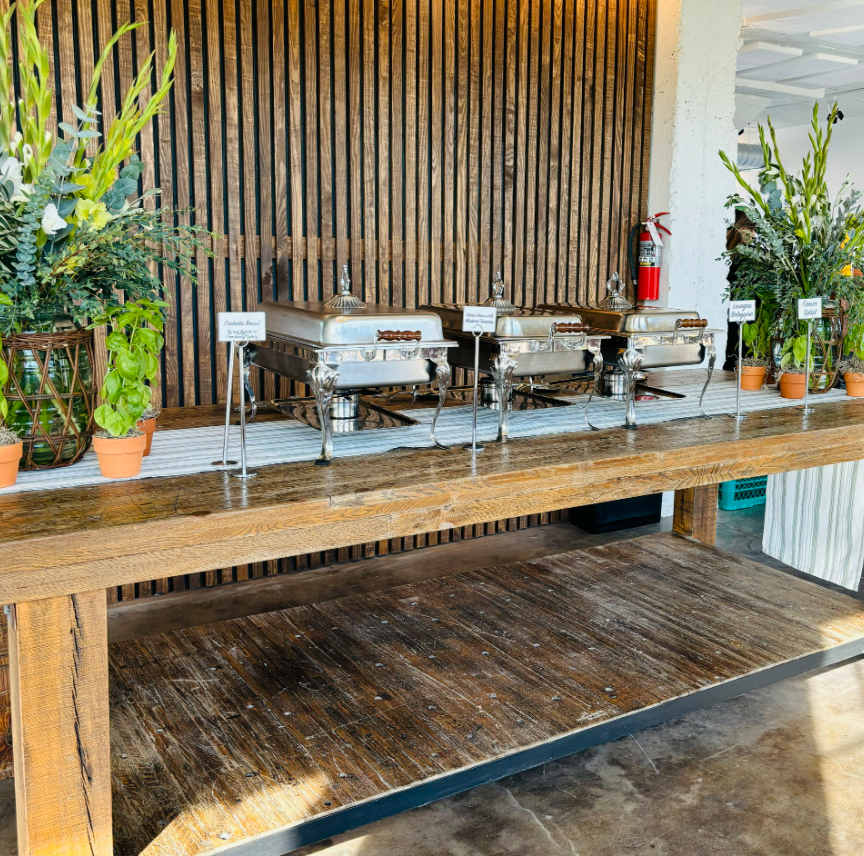 Buffet table with three chafing dishes, potted plants, and a fire extinguisher against a wooden slat wall.