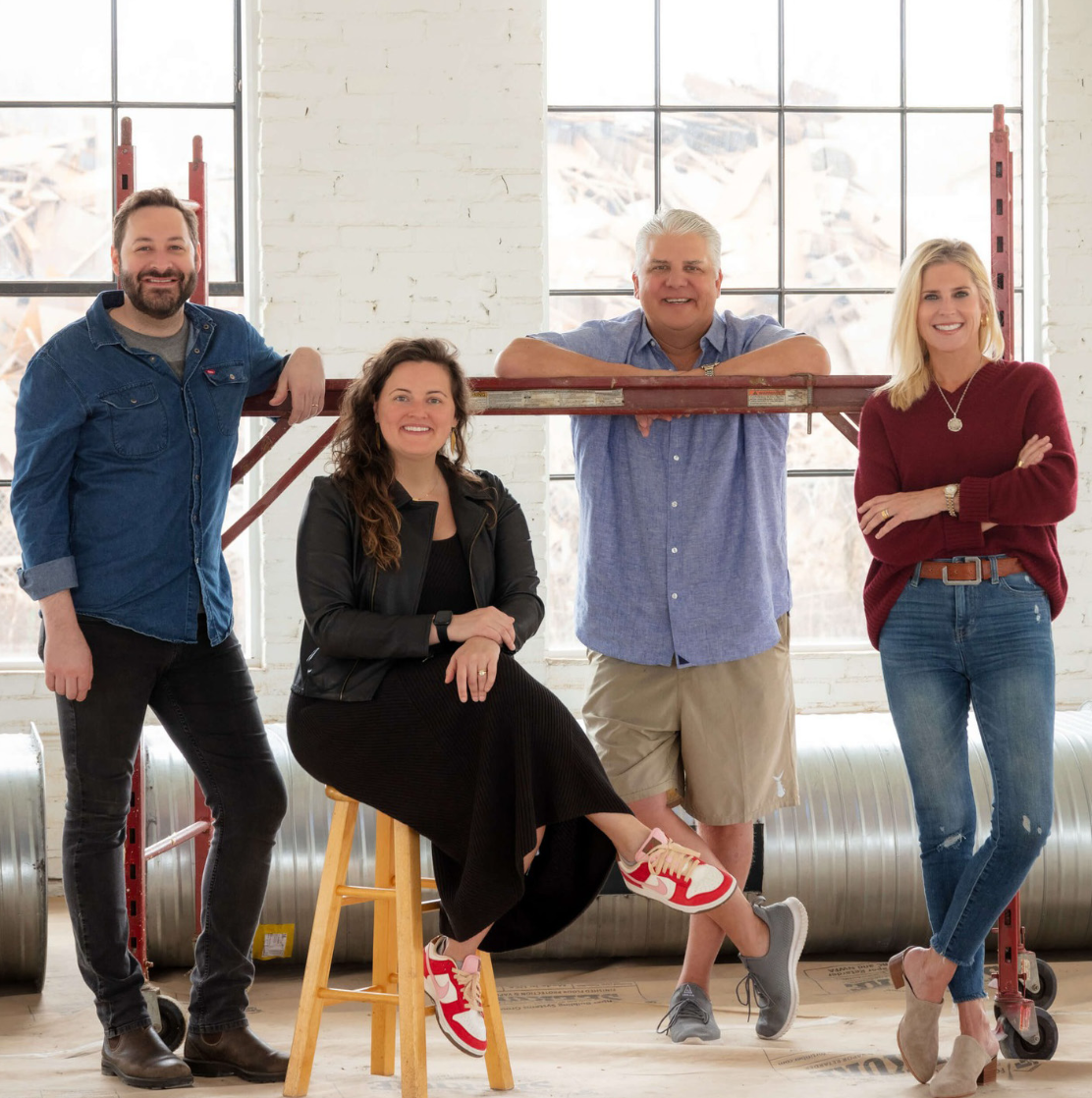 The owners of Eastside Scrapyard pose in a bright industrial-style space with large windows, white brick walls, and construction scaffolding.