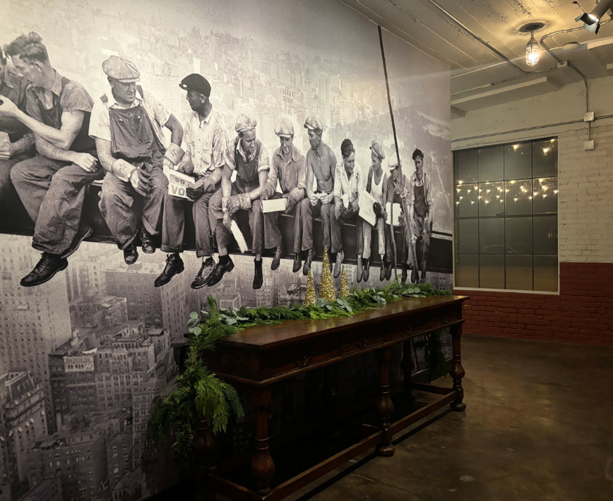 Black and white mural of historical factory workers sitting on a ledge, with a decorated table and greenery below, in an industrial-style room.