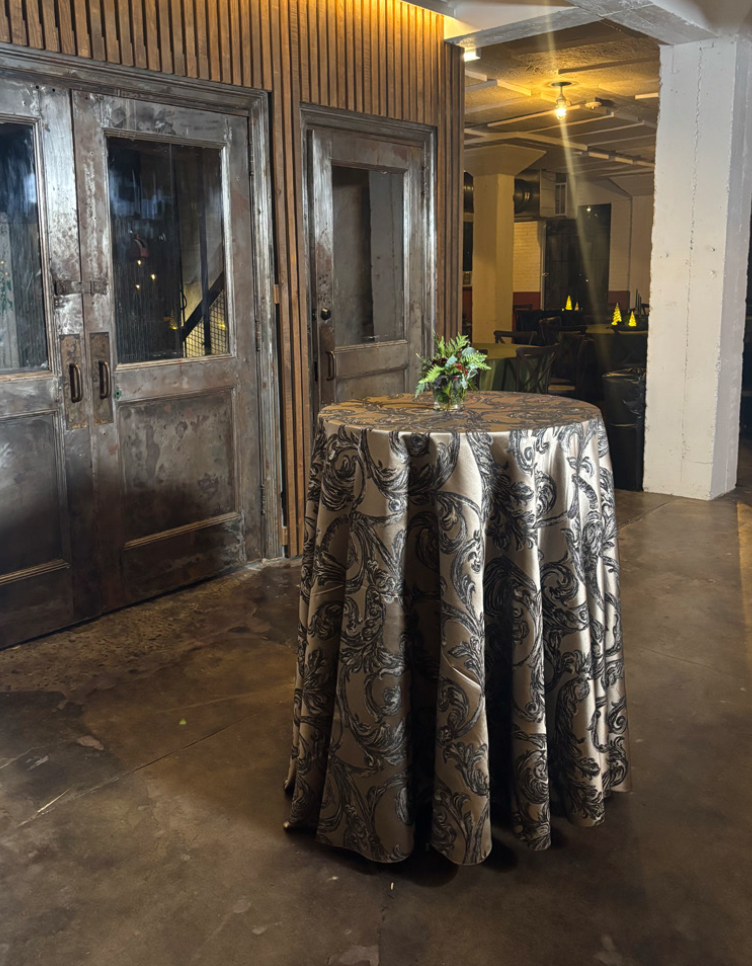A round table covered with a patterned cloth and a small floral centerpiece in a dimly lit room with wooden and concrete walls and a double door.