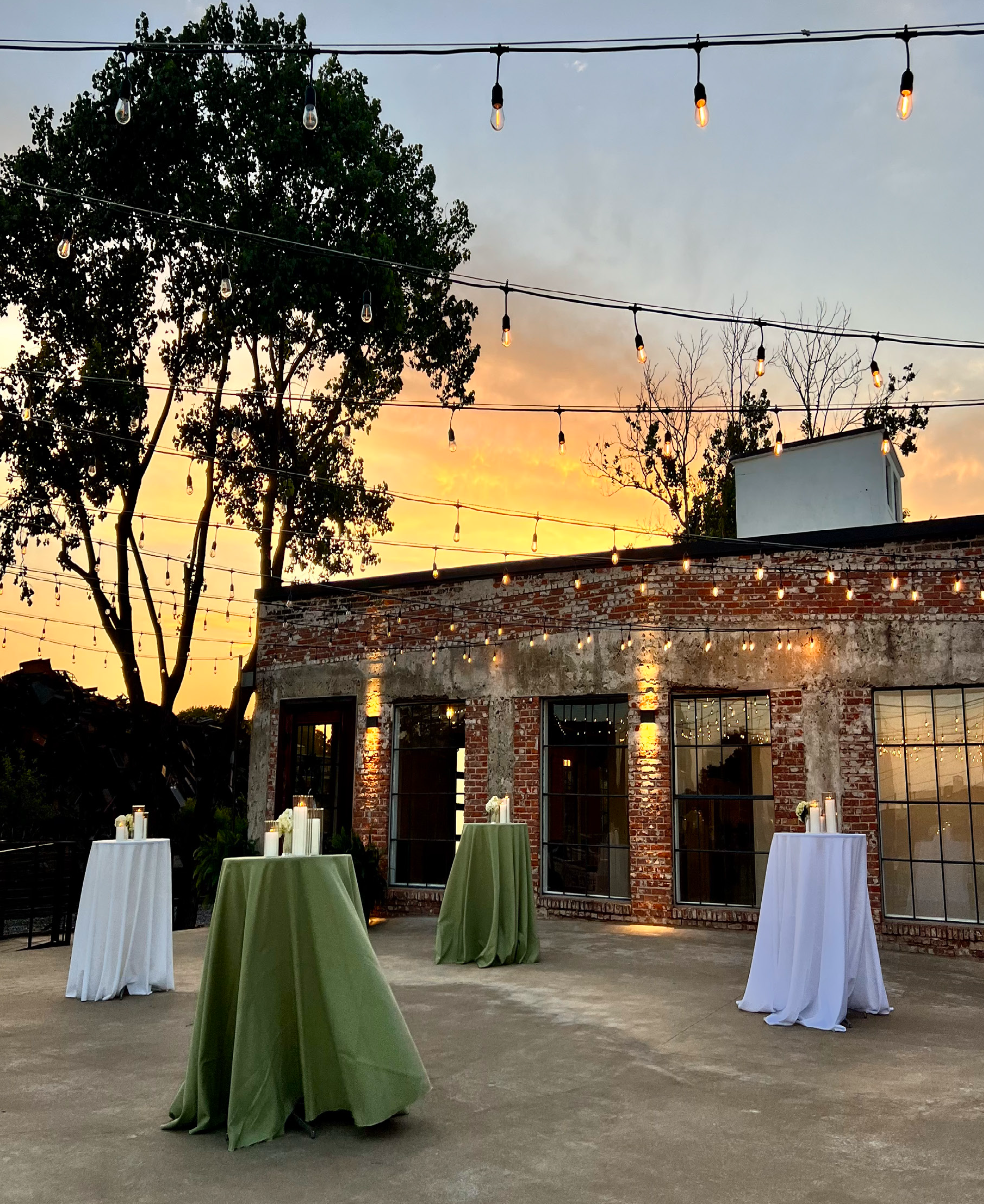 Outdoor evening setting with string lights overhead, brick building with large windows in the background, and four tall tables draped with white and green tablecloths, lit candles on each table, at sunset.