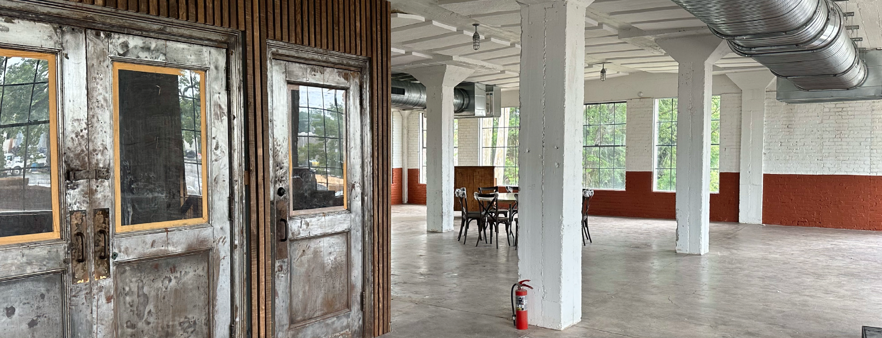The image shows an industrial-style interior space with large windows, white brick walls with a red lower half, and white support columns. There are exposed duct pipes on the ceiling and a fire extinguisher on the floor. A rustic wooden door and some metal chairs and a table are visible in the background.