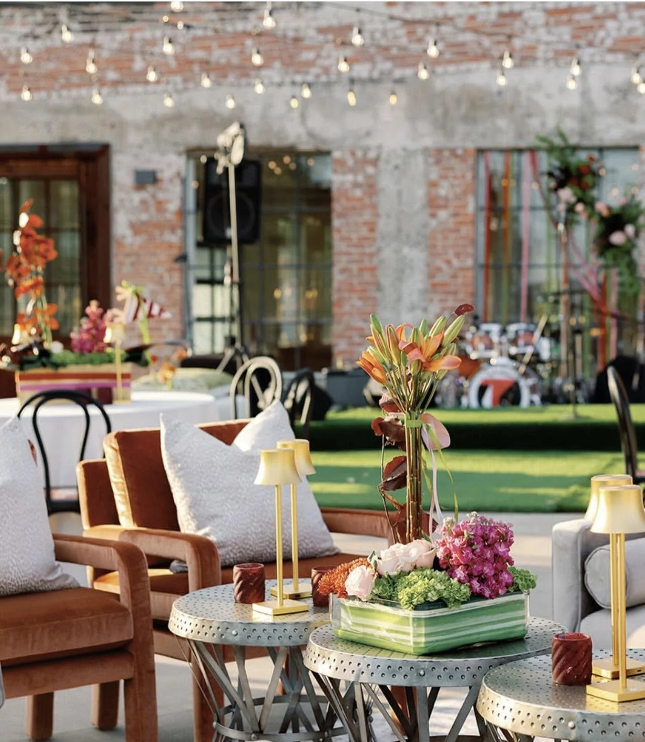 Decorated outdoor patio with floral centerpieces, tables, and chairs, set for an event against a brick wall with string lights.