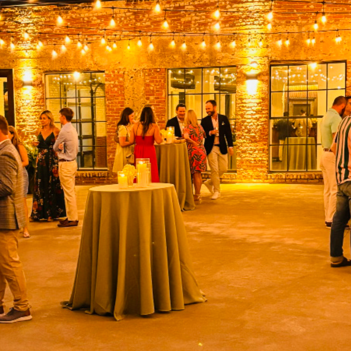 People socializing at an indoor evening event with warm lighting, brick walls, and large windows, standing around high tables with drinks and candles.