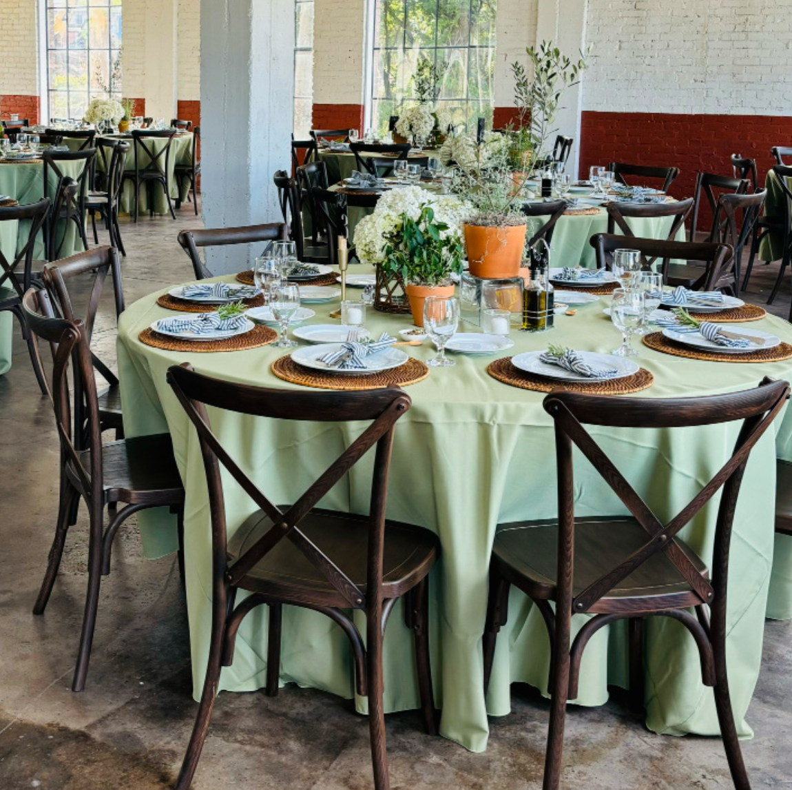Set dining table with green tablecloth, white plates, striped napkins, glassware, and floral centerpiece in a bright, industrial-style venue with large windows and exposed brick walls.