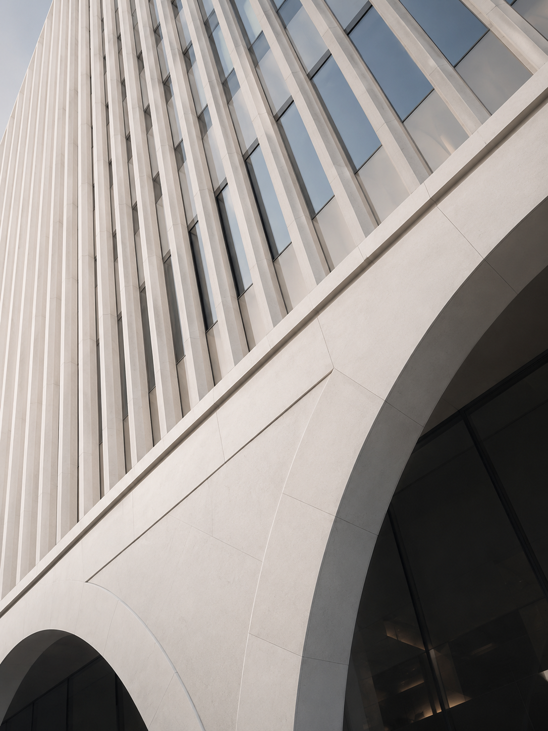 Low-angle view of a modern high-rise building with vertical white panels and large reflective windows, featuring an arch at the base.