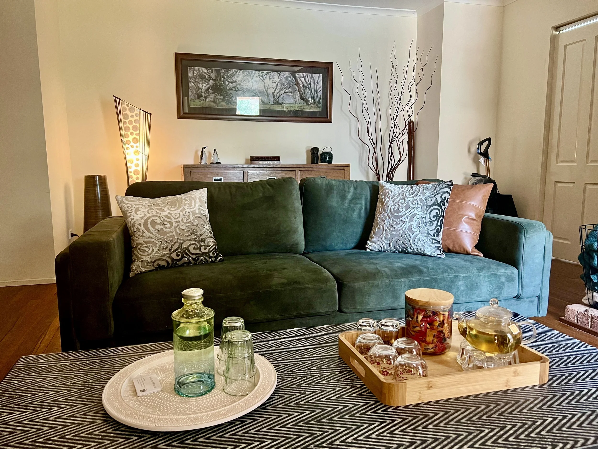 Living room with green sofa, decorative pillows, a coffee table with glasses and containers, a wooden sideboard with accessories, framed landscape picture on the wall, floor lamp, and tall decorative branches.
