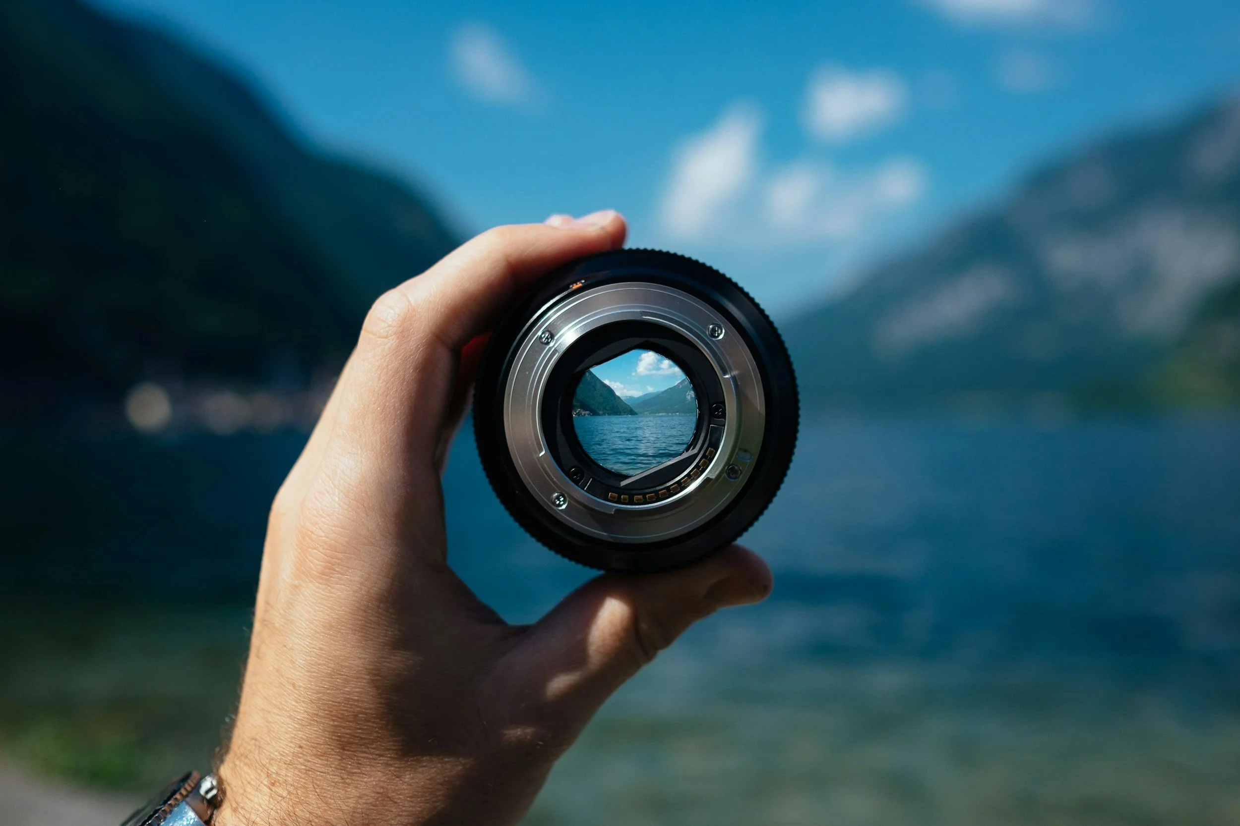 A person holding a camera lens in front of a scenic landscape featuring a lake surrounded by mountains and blue sky with clouds.