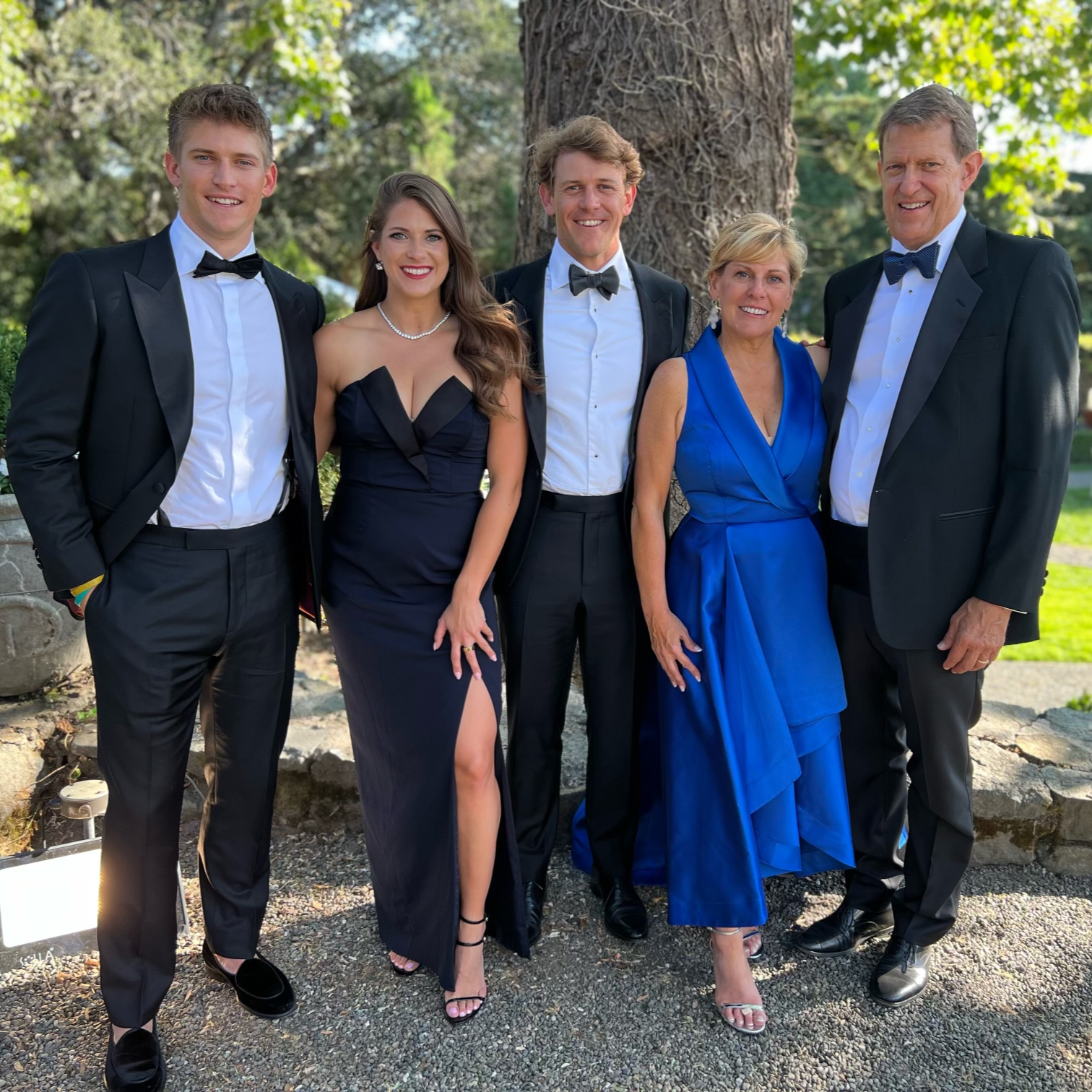 Ginny Merrifield and family dressed in formal attire, standing outdoors next to a large tree, smiling at the camera.