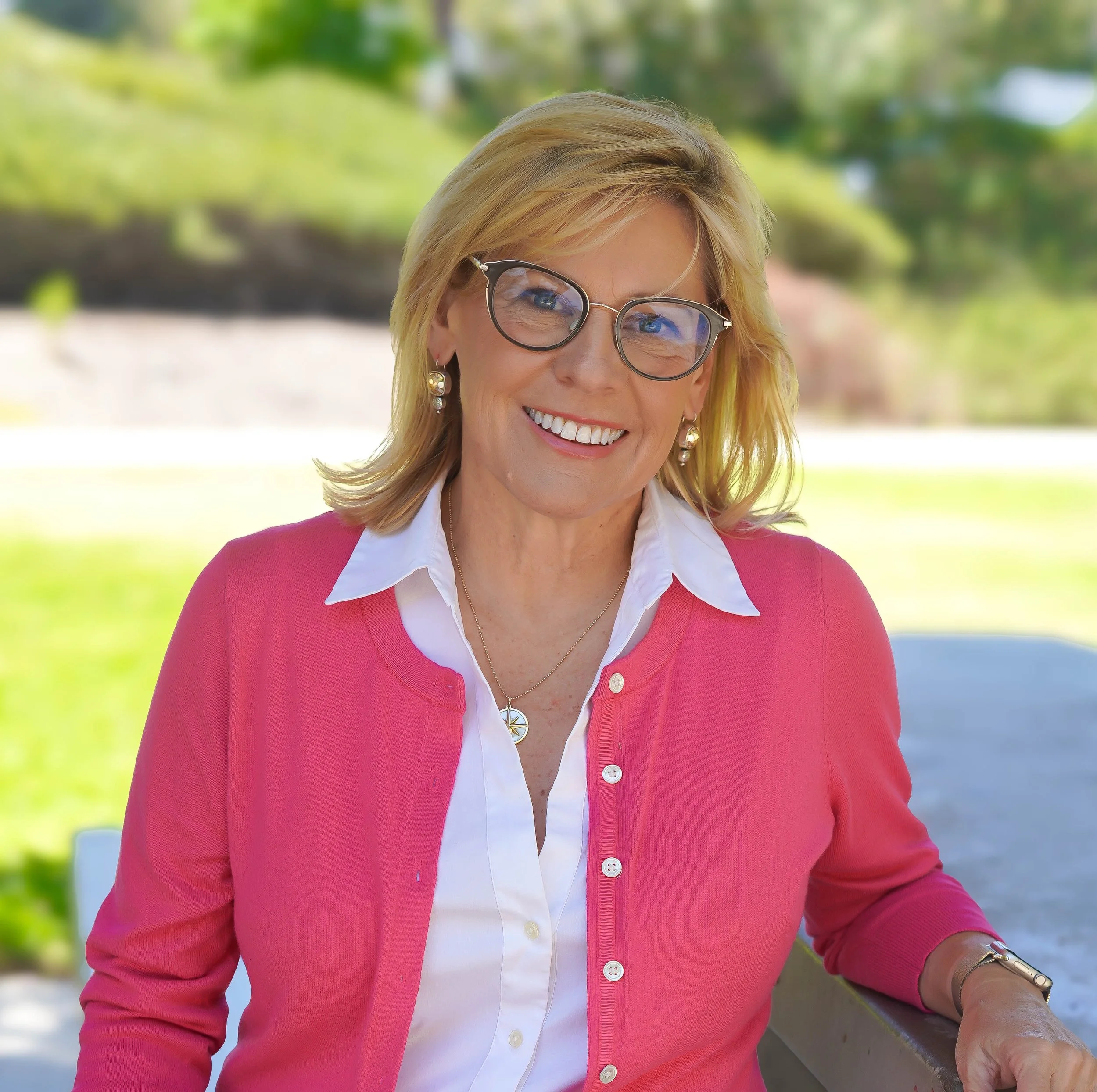 Close-up of a woman with blonde hair, wearing glasses, earrings, a white shirt, and a pink cardigan, smiling outdoors with blurred greenery in the background.