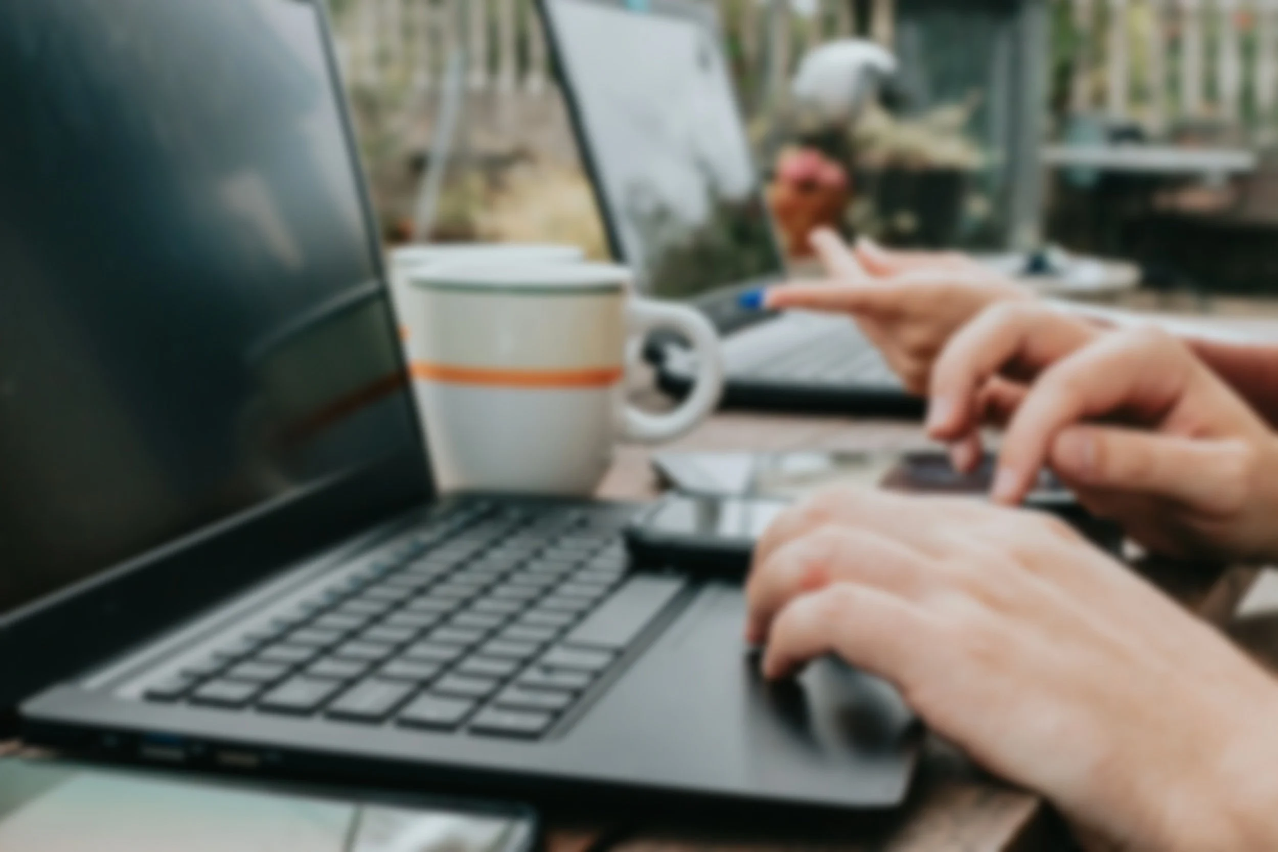 Close-up of of several hands typing on laptop keyboards at an outside table, with a mug nearby and a blurred background.