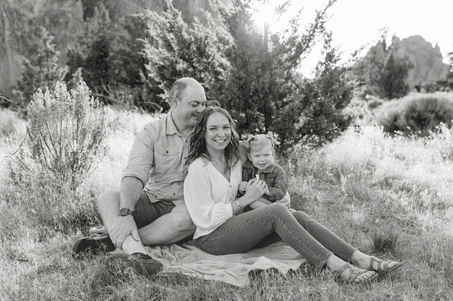 A black and white image of Katelin Williams' family of three sitting on a blanket outdoors among trees and plants at Smith Rock State Park,  smiling at the camera.