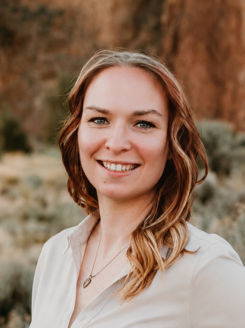 Smiling person with shoulder-length light brown and blonde hair wearing a light-colored blouse and necklace, posing outdoors with a blurred natural background.