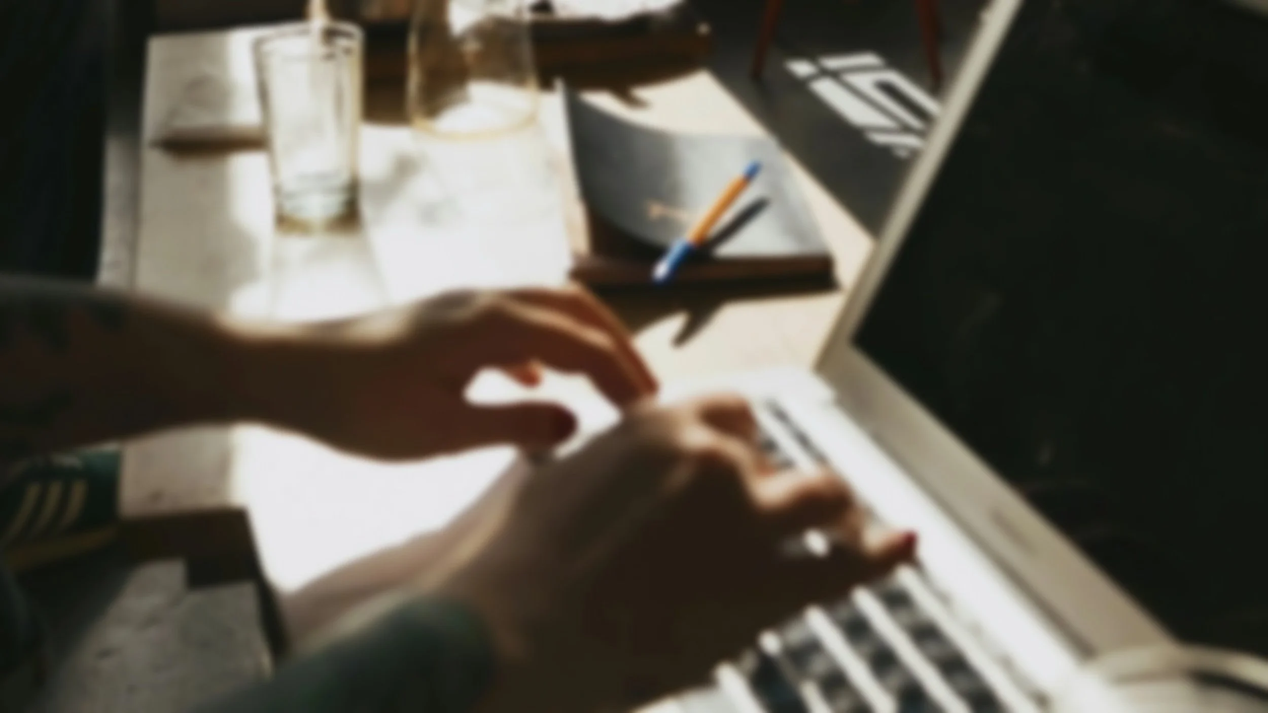 Blurred image of a person's hands typing on the keyboard with a notebook, pen, and glasses on a desk, sunlight casting shadows.