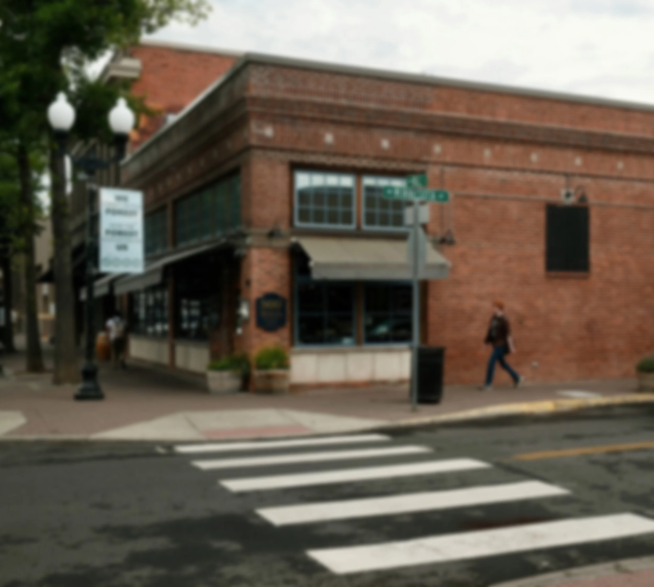 Blurry image of a city street scene in downtown Bend, Oregon with a brick building, street signs, a pedestrian crossing, and a person walking on the sidewalk.