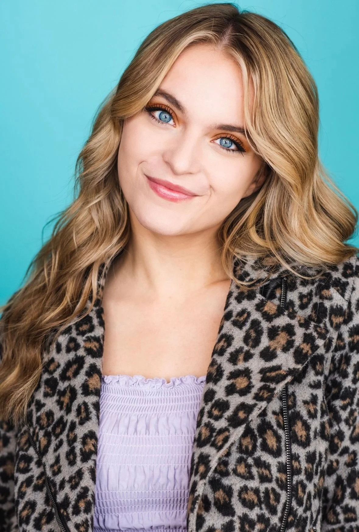 A young woman with long, wavy blonde hair and blue eyes, smiling, wearing a leopard print jacket over a light lavender top, against a solid turquoise background.