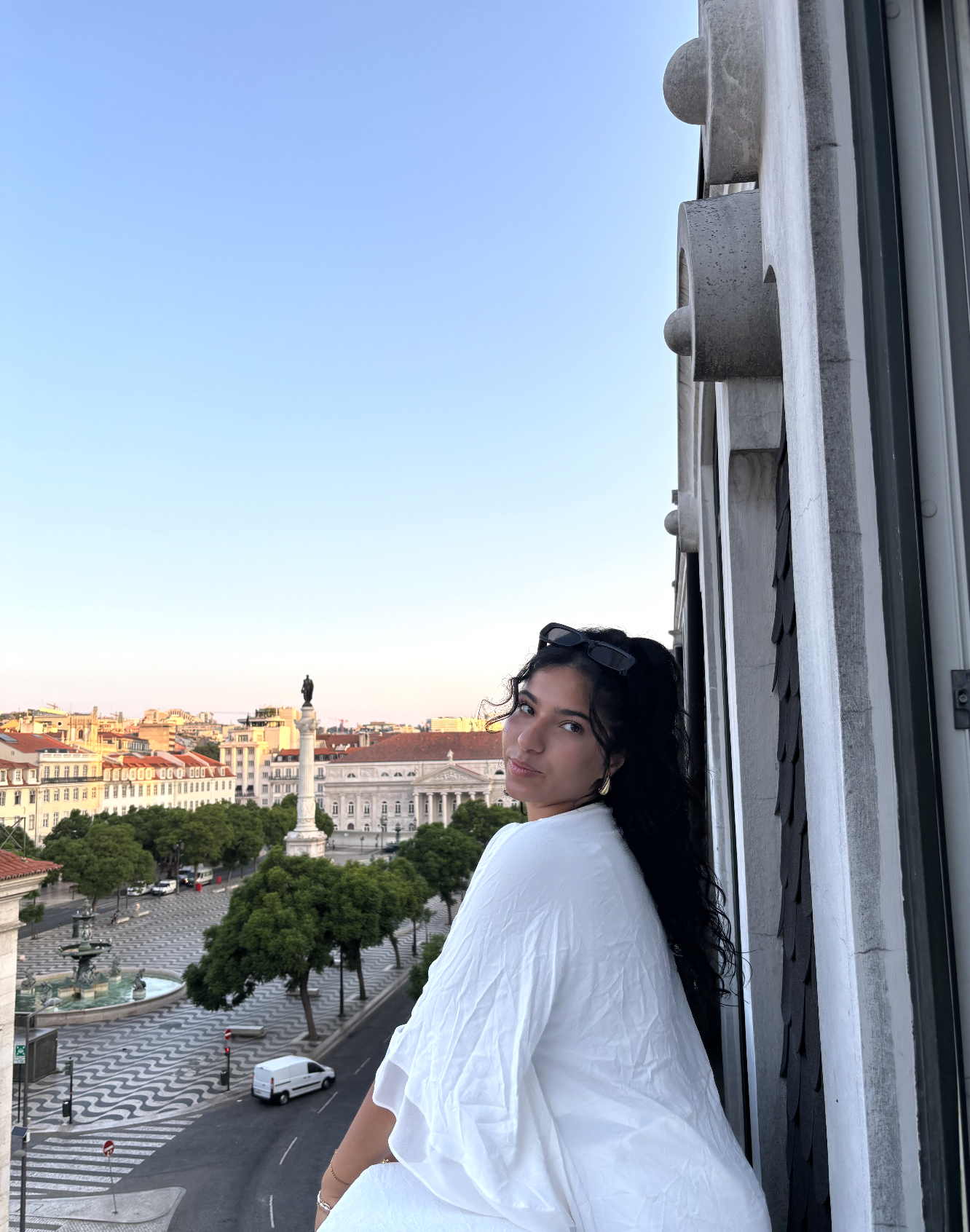 A woman with dark curly hair and sunglasses on her head wearing a white dress sits on a balcony overlooking a city square with a statue and historic buildings during sunset.