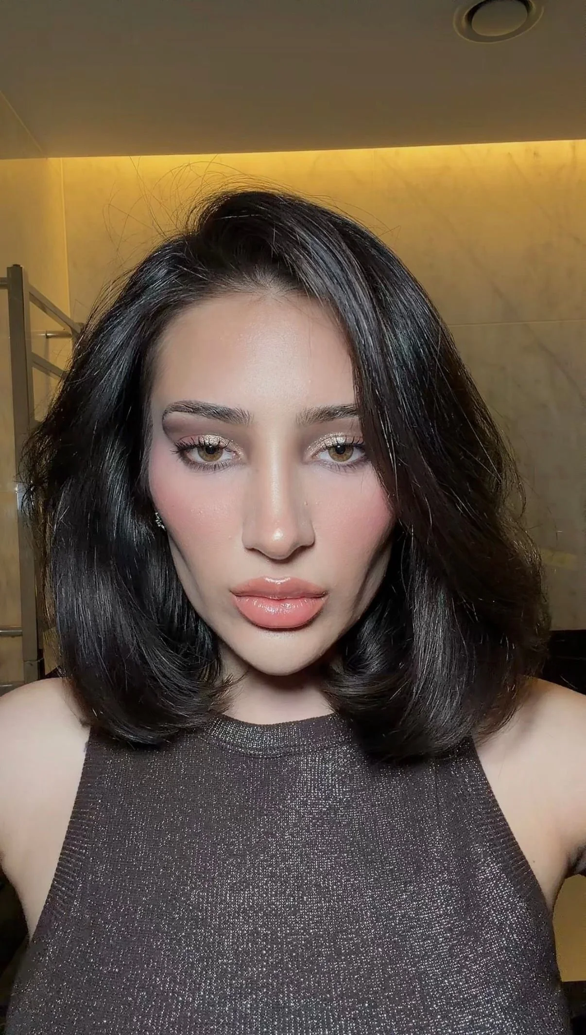 A close-up of a woman with shoulder-length dark hair, wearing a sleeveless, sparkly dark top, with makeup including nude lips and shimmery eye makeup, standing in front of a marble wall and yellow lighting.
