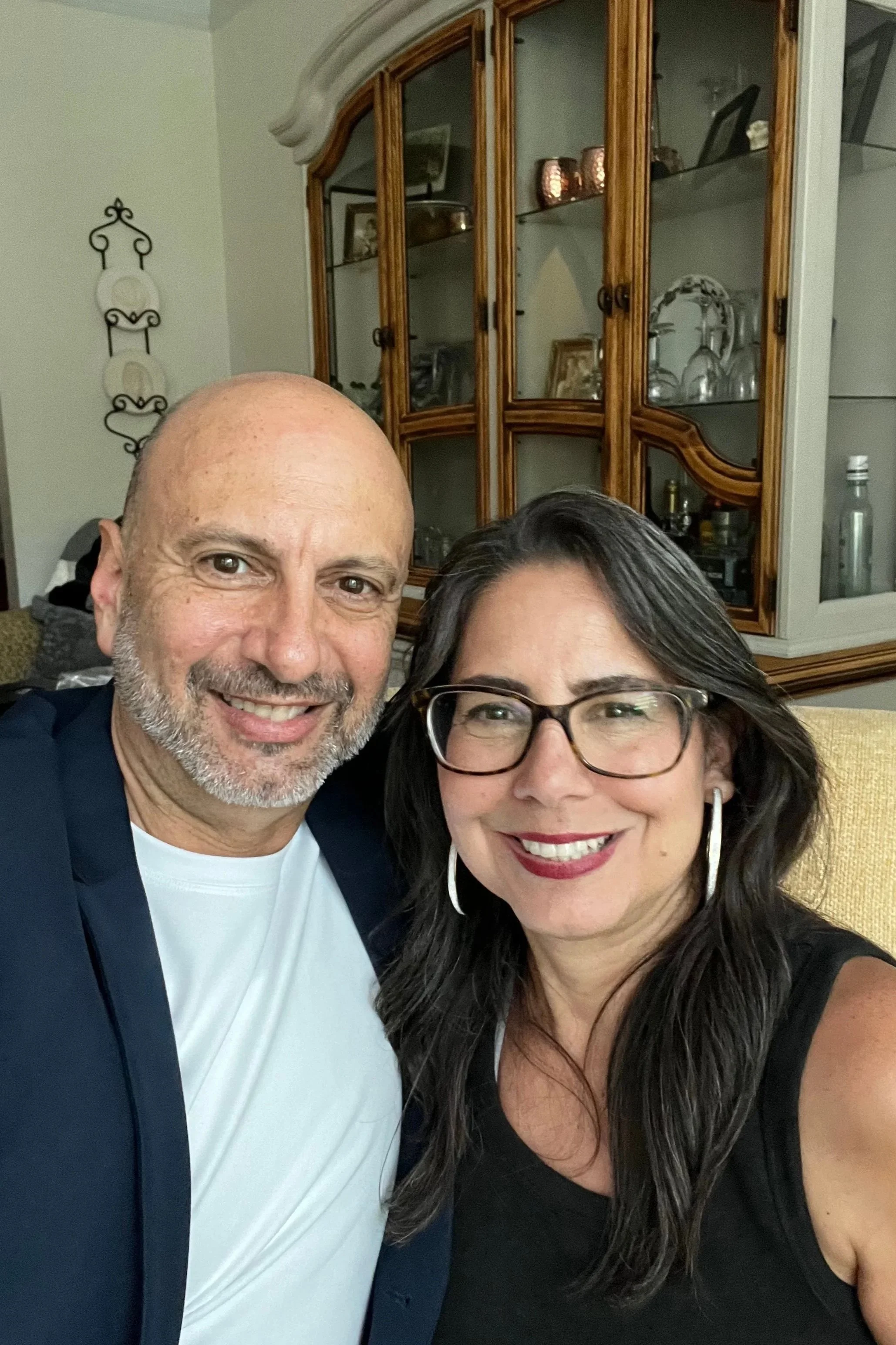 Close-up selfie of a man with a bald head and beard, wearing a white shirt and dark blazer, and a woman with long dark hair, glasses, and earrings, smiling in a living room setting with a wooden cabinet in the background.