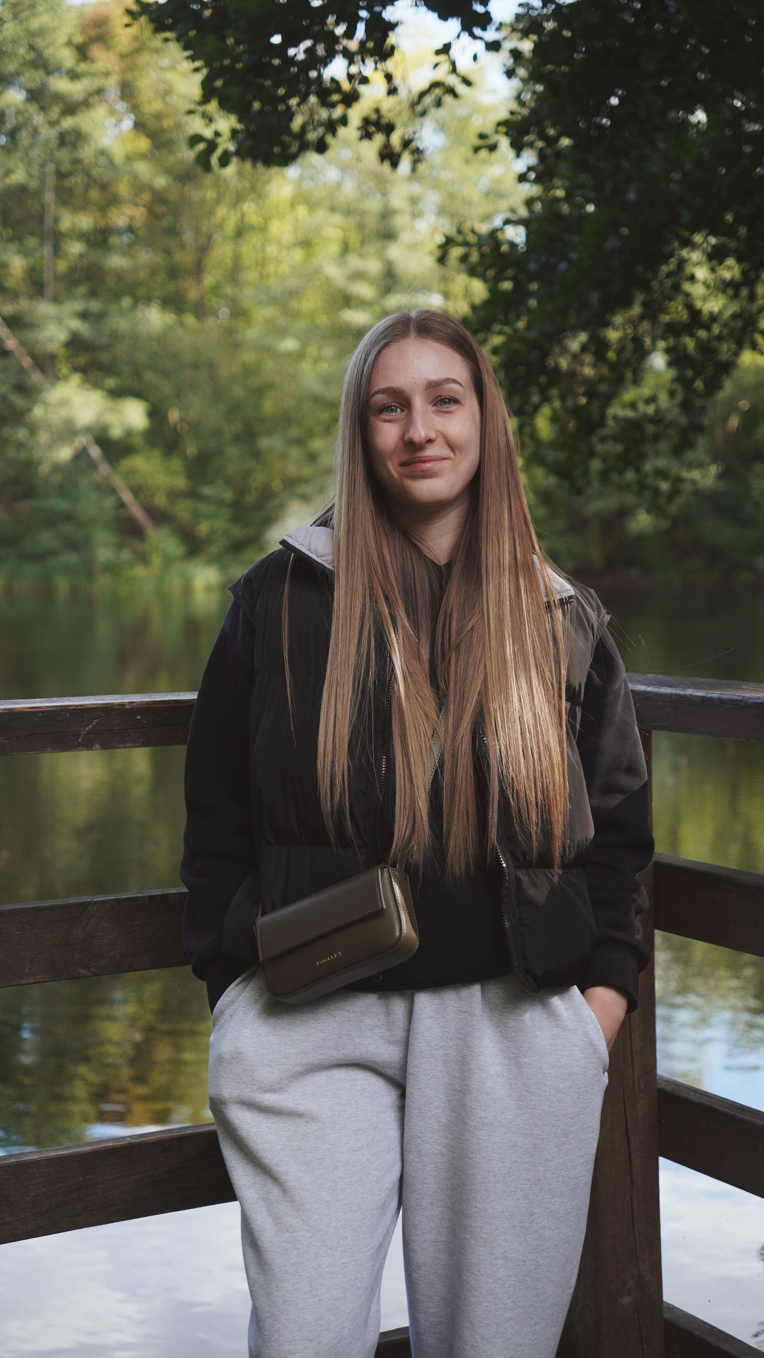 A young woman with long, straight brown hair standing on a wooden bridge in a lush, green park by a river, wearing a black jacket, gray sweatpants, and a small olive green purse.