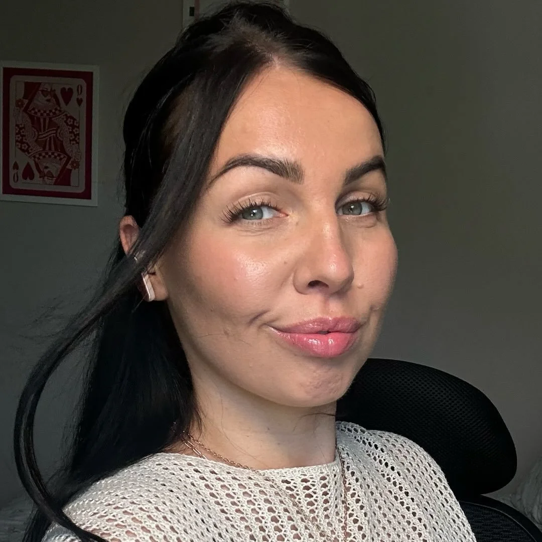A woman with dark hair, light skin, and blue eyes, smiling slightly, wearing a beige knitted top and a delicate necklace, sitting in front of a gray background with a black chair behind her.