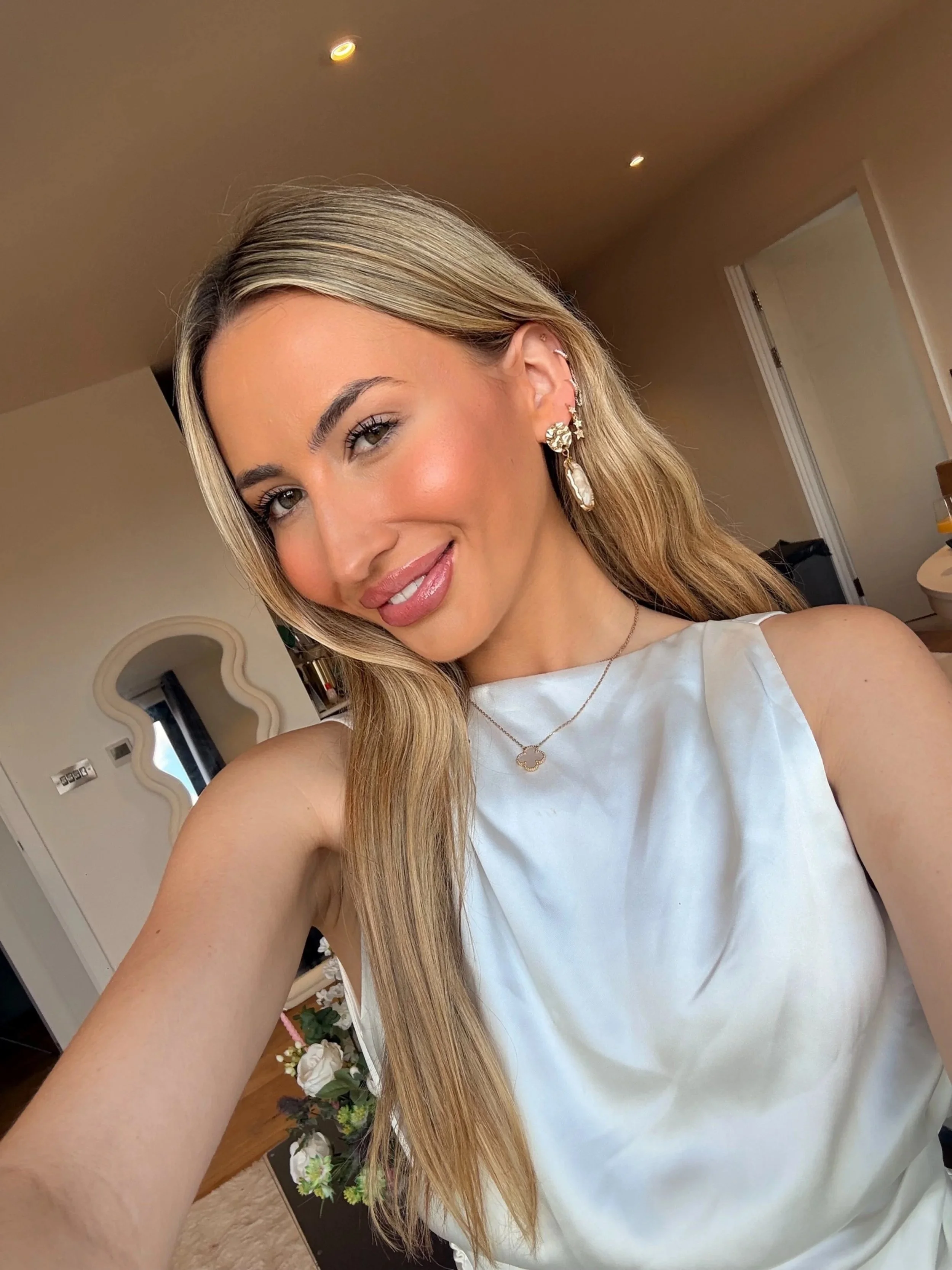A young woman with long blonde hair taking a selfie indoors, smiling softly, wearing earrings and a white sleeveless top.