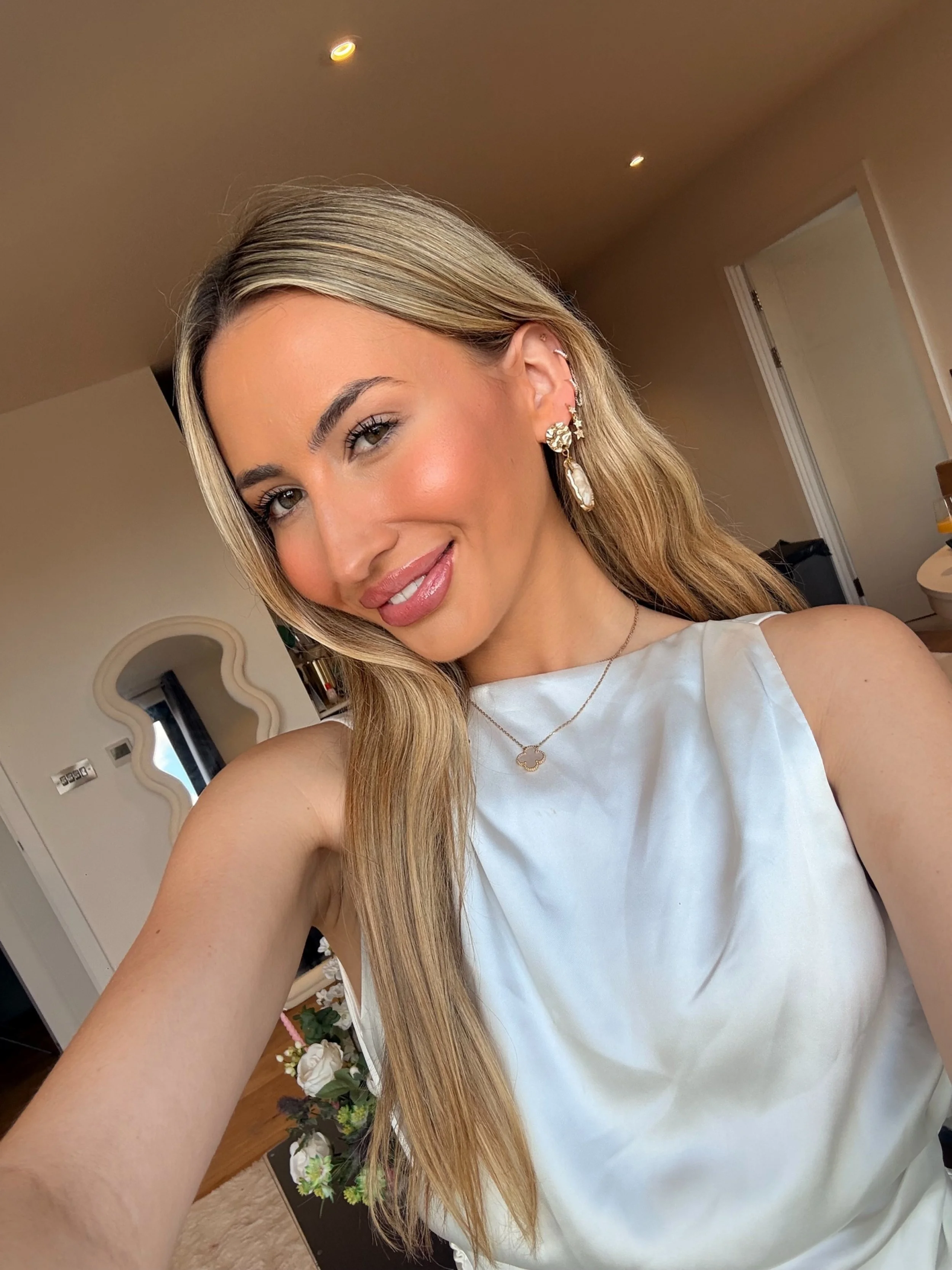 A woman with long blonde hair taking a selfie in a room. She is smiling, wearing large earrings, a white sleeveless top, and a delicate necklace. The background includes a doorway, a mirror, and a flower arrangement.