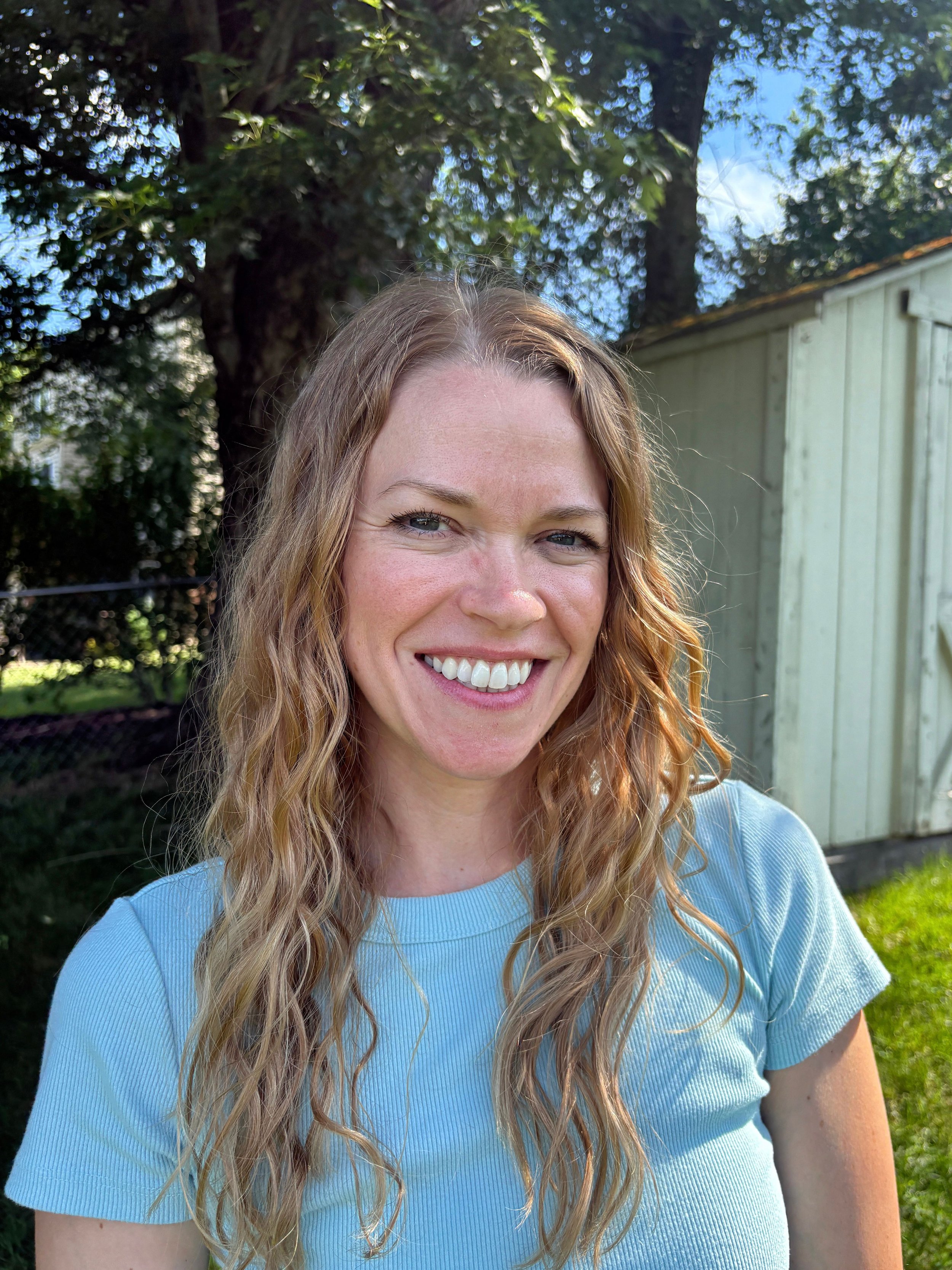Close-up of a woman with long, curly red hair smiling outdoors with green trees and a light yellow shed in the background.