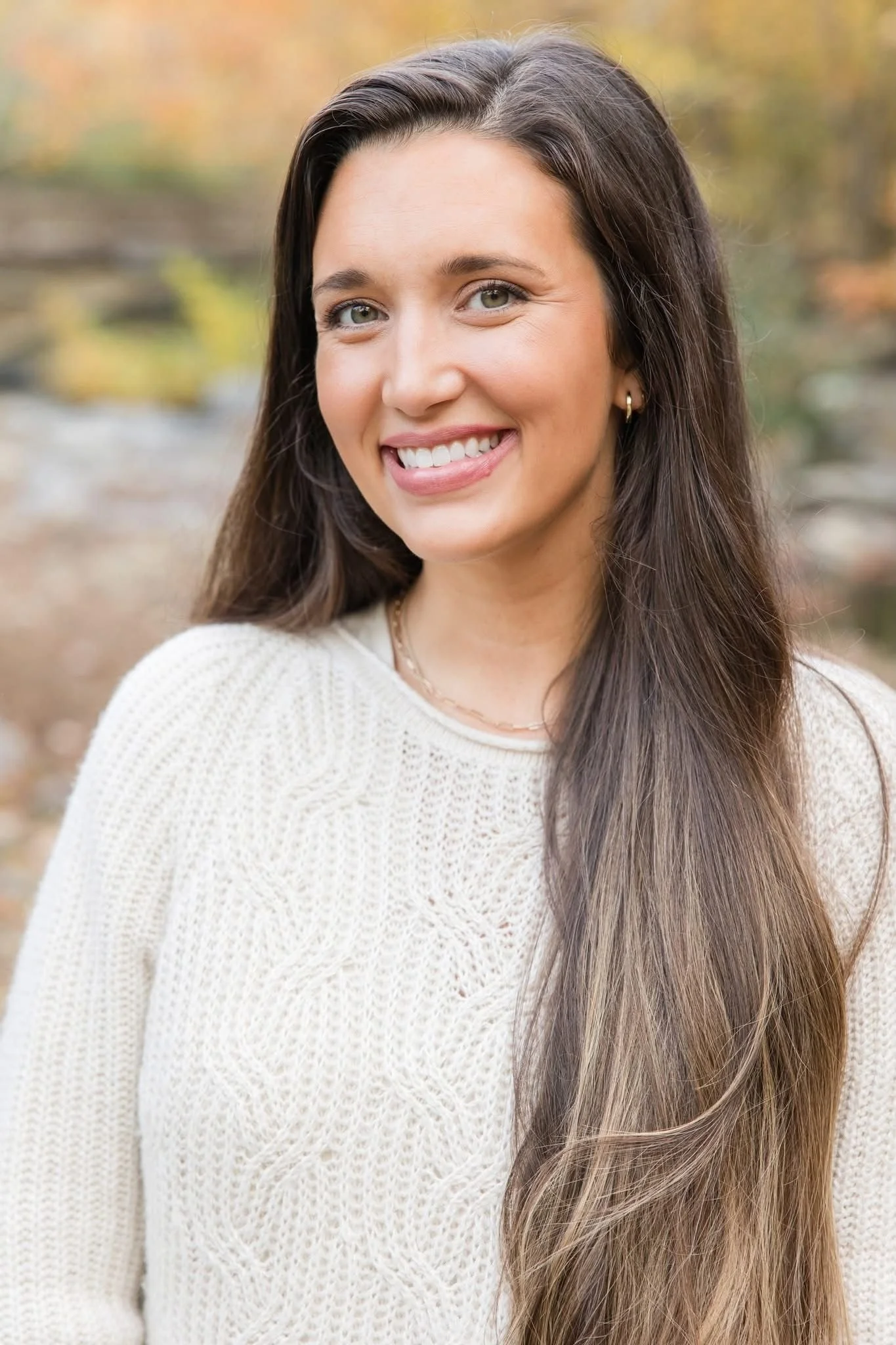 A woman with long brown hair and light skin, smiling, outdoors with autumn-colored blurred background, wearing a cream knit sweater.