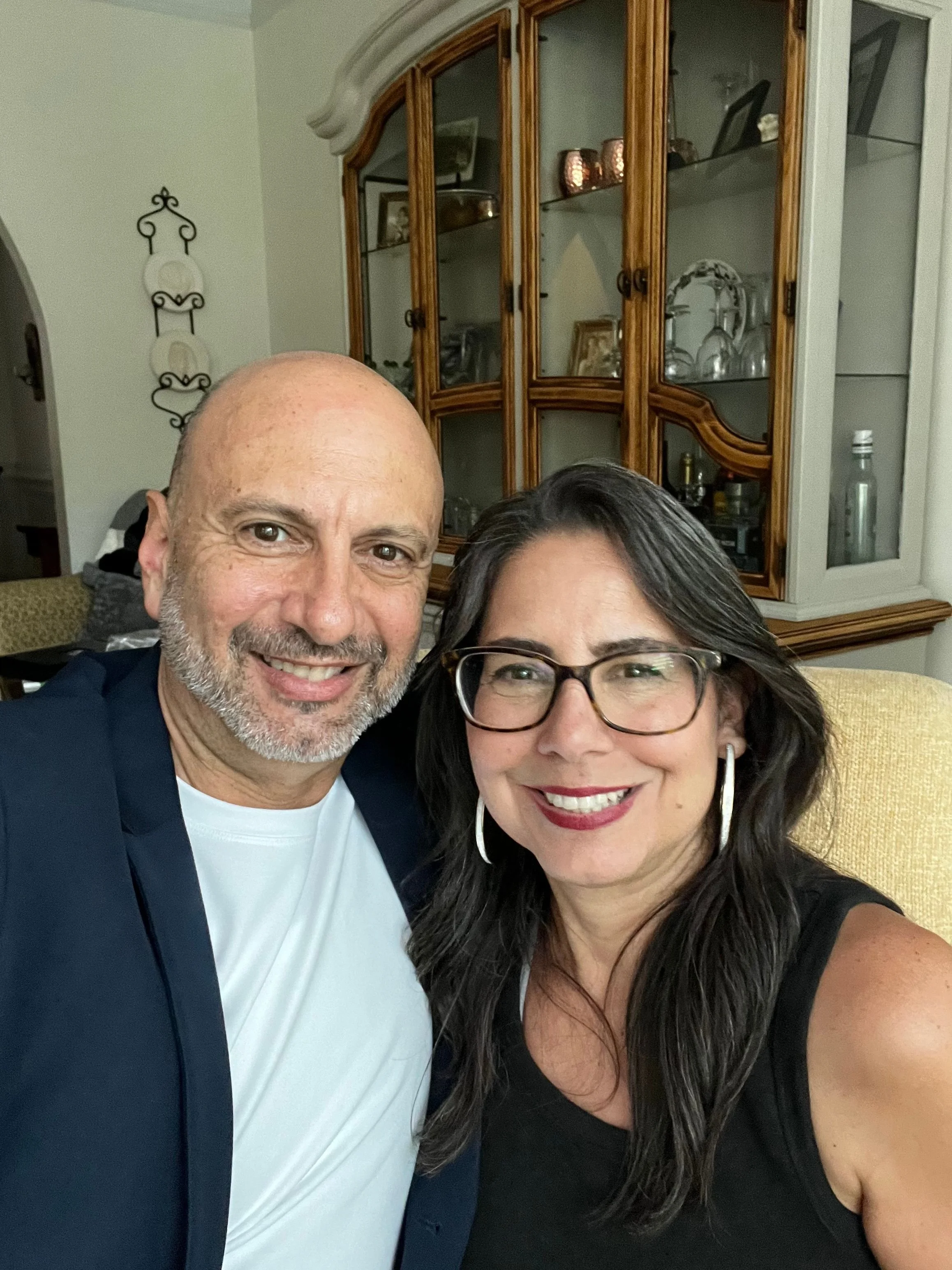 A smiling man and woman taking a selfie together indoors near a wooden display cabinet.