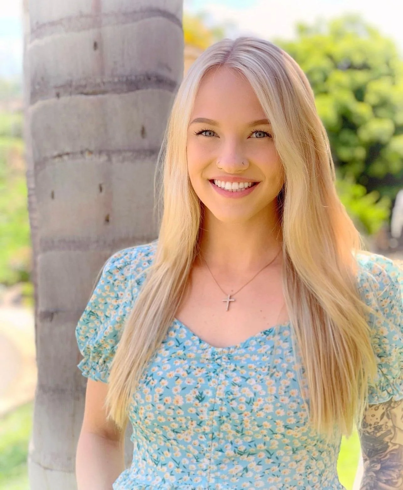 Young woman with long blonde hair, smiling, wearing a blue floral dress and cross necklace outdoors near a palm tree with a green leafy background.