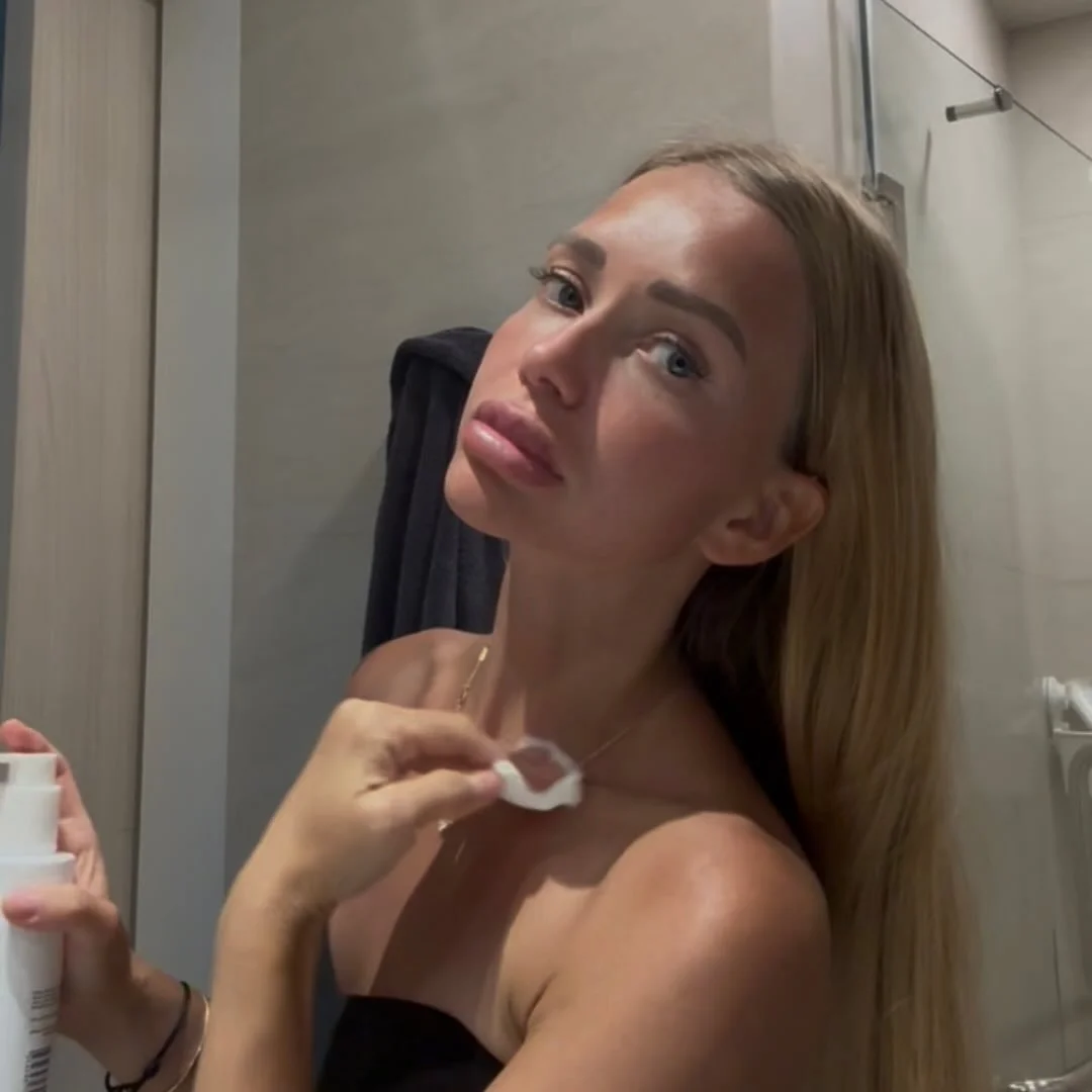 A young woman with long blonde hair and blue eyes is in a bathroom, holding a cotton pad near her neck, and looking into a mirror.
