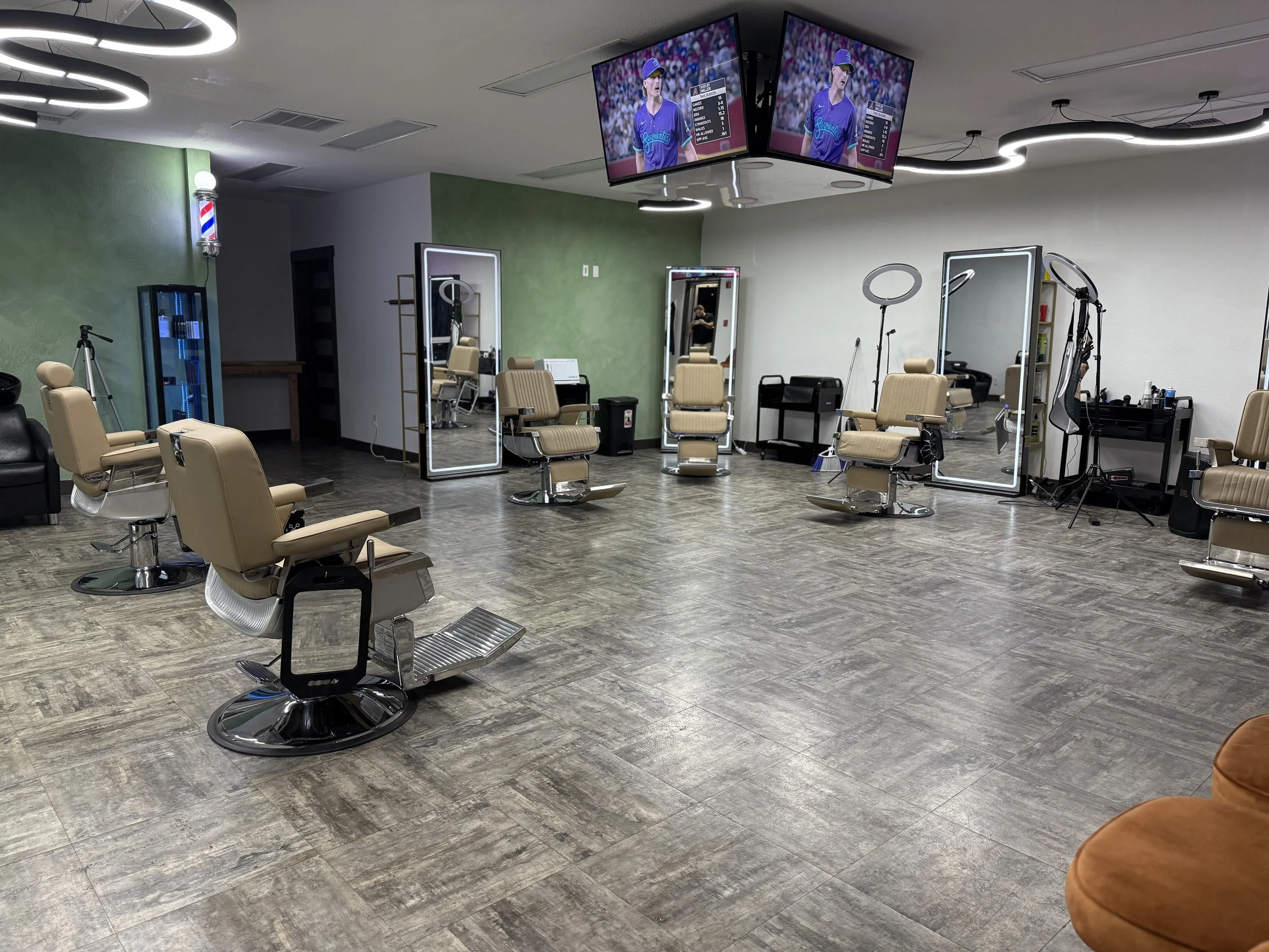 A hair salon features multiple styling chairs and mirrors, with a large overhead TV displaying a sports game and a green accent wall.