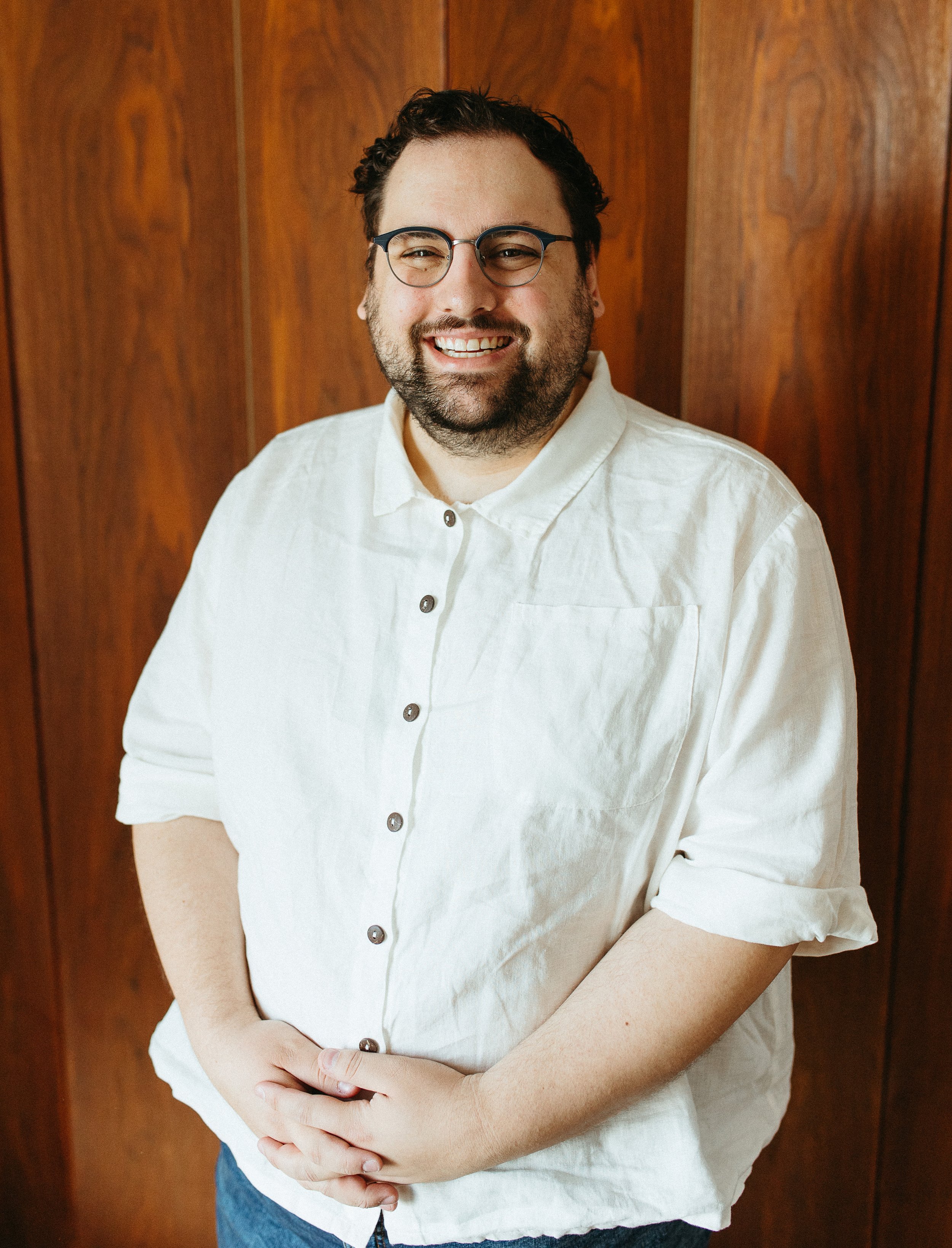 A smiling man wearing glasses and a white button-up shirt stands against a wooden wall background, with his hands clasped in front of him.