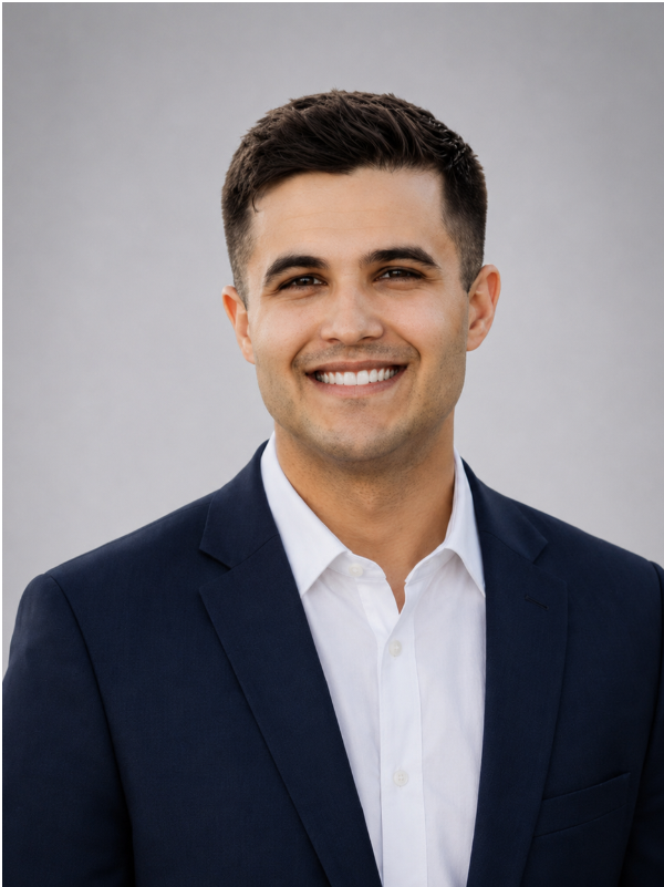 A young man with short dark hair, wearing a navy suit jacket and a white dress shirt, smiling at the camera against a plain gray background.