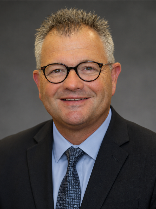 Portrait of a middle-aged Caucasian man with short, spiky gray hair, wearing round black glasses, a dark suit, light blue shirt, and patterned tie, smiling against a gray background.