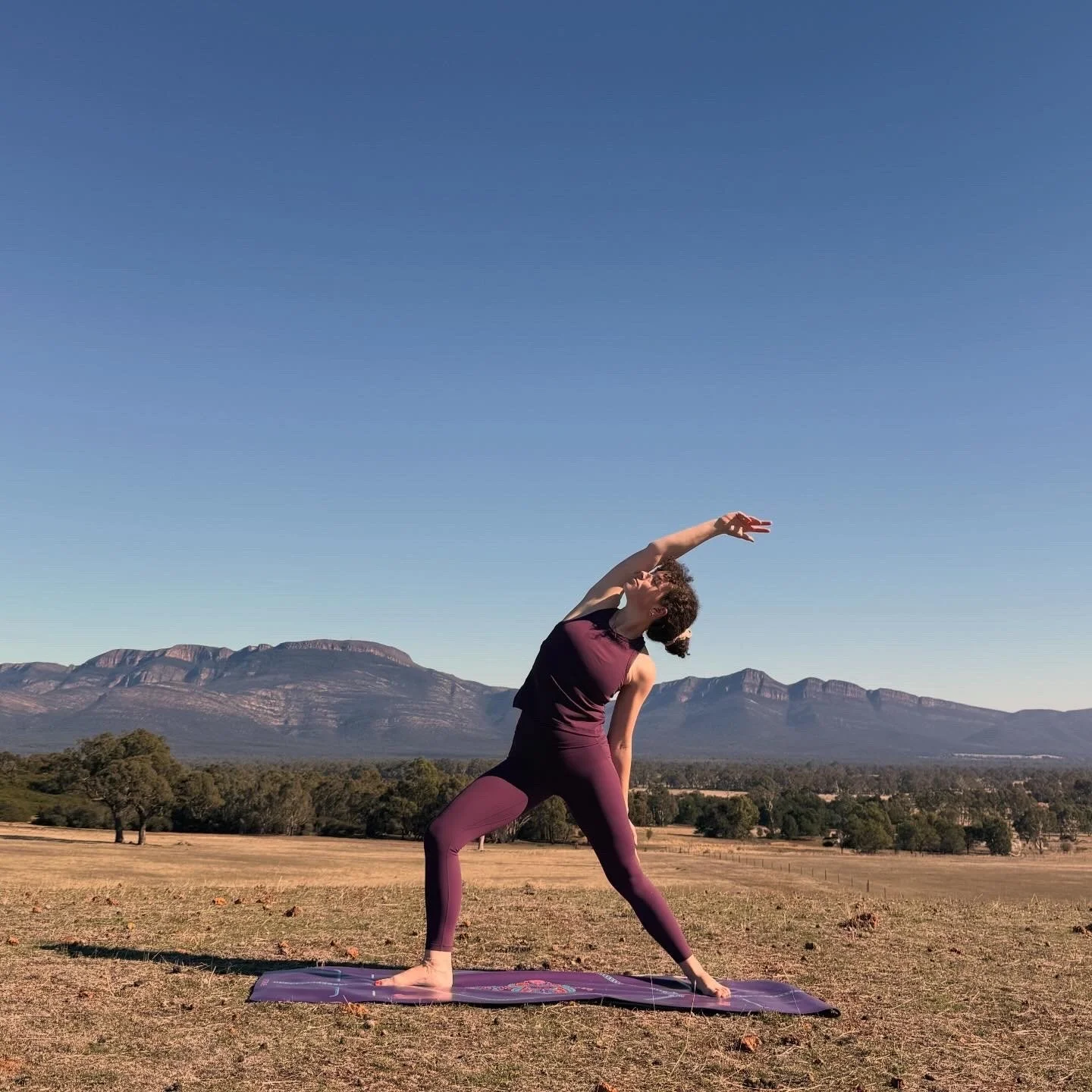 A woman practicing yoga outdoors on a purple mat in a field with mountains in the background under a clear blue sky.