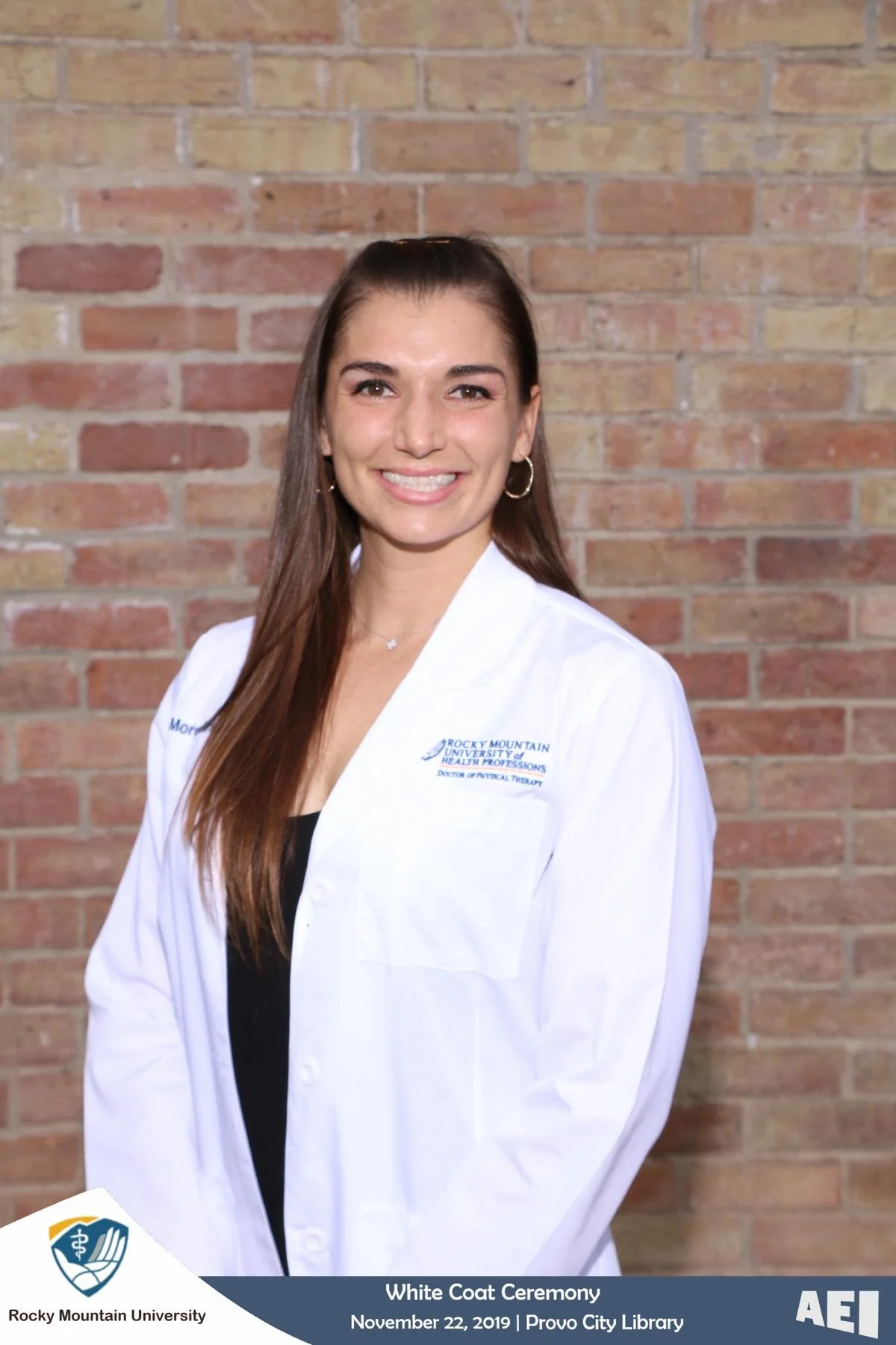 A woman with long brown hair wearing a white coat and earrings, smiling against a brick wall, during Rocky Mountain University White Coat Ceremony on November 22, 2019 at Provo City Library.