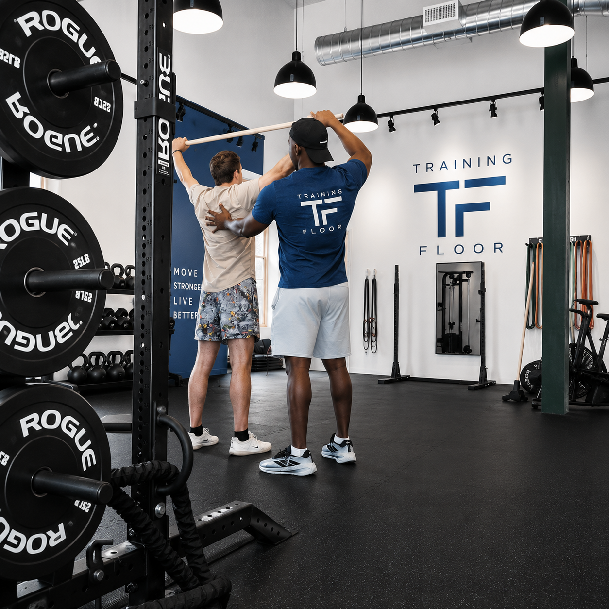 A fitness trainer assisting a young man in a gym with a stretching exercise. The gym has black rubber flooring, weight plates, kettlebells, and resistance bands. The background features a blue wall with the words "MOVE STRONGER LIVE BETTER" and a logo that says "Training Floor."