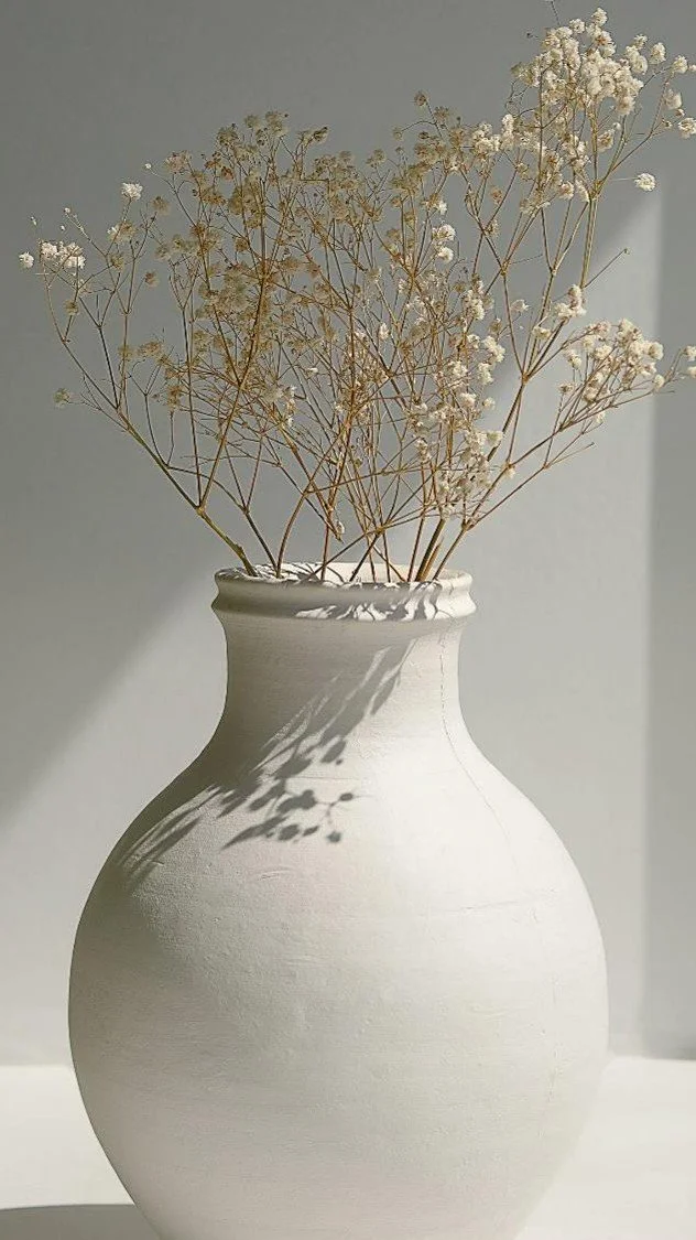 A white ceramic vase holding dried white baby's breath flowers, casting shadows on a light gray wall.