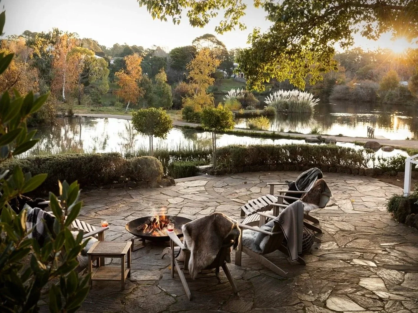 A cozy patio with a fire pit, lounge chairs with blankets, overlooking a pond with trees and shrubs at sunset.