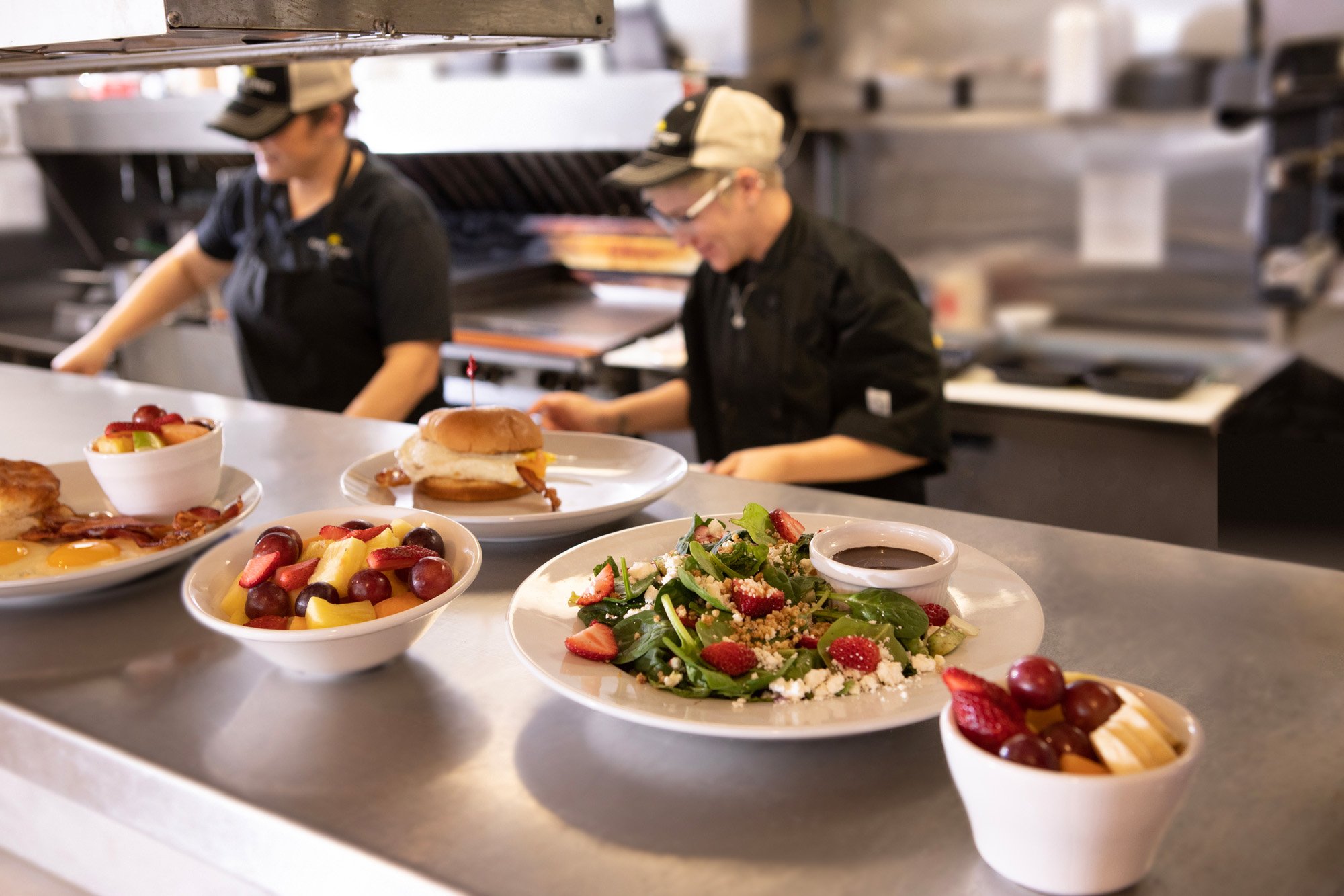 Assorted breakfast dishes including bowls of fruit salad with grapes, pineapple, and strawberries, a vegetable salad with strawberries, spinach, cheese, and dressing, grilled breakfast sandwiches with eggs, cheese, and bacon, and a bowl of cereal or sauce, on a counter in a commercial kitchen with two chefs working in the background.