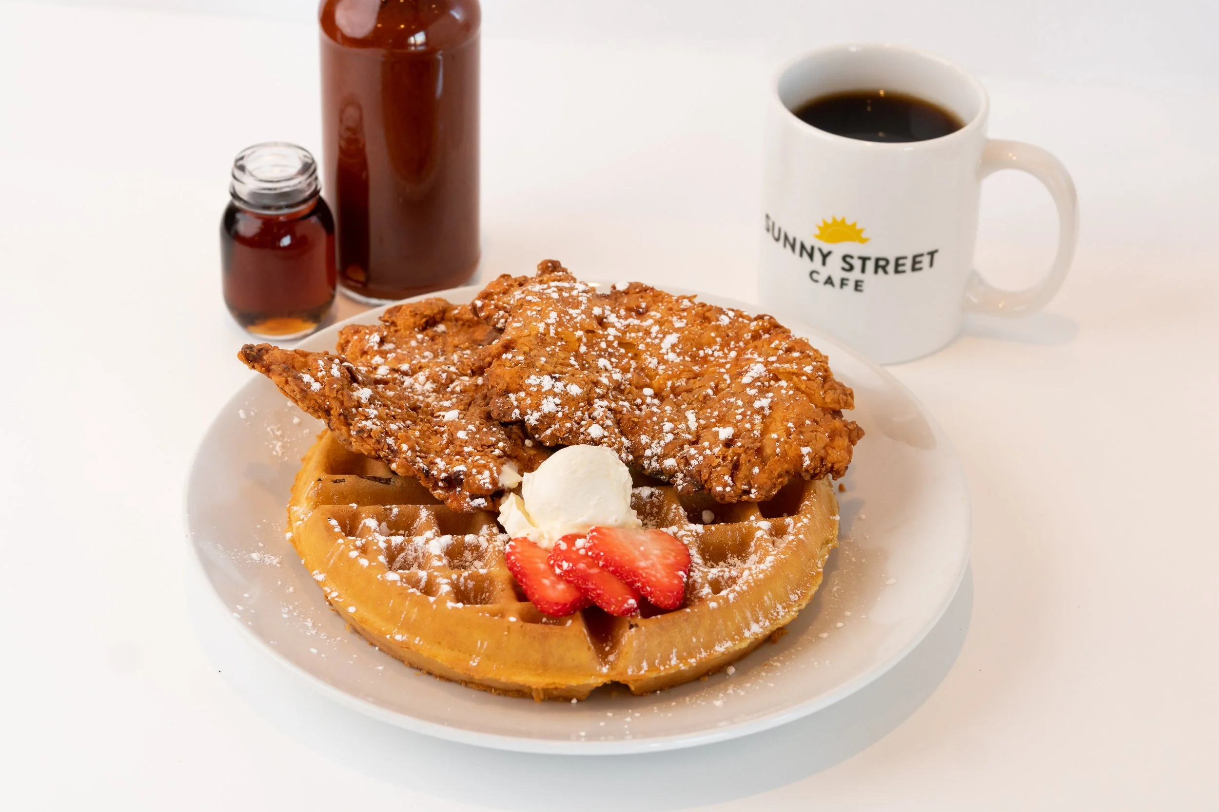 A plate with a waffle topped with strawberries, whipped cream, a fried chicken fillet, and powdered sugar, served with a dollop of butter. In the background, there's a coffee mug with the logo 'Sunny Street Cafe' and a bottle of syrup.