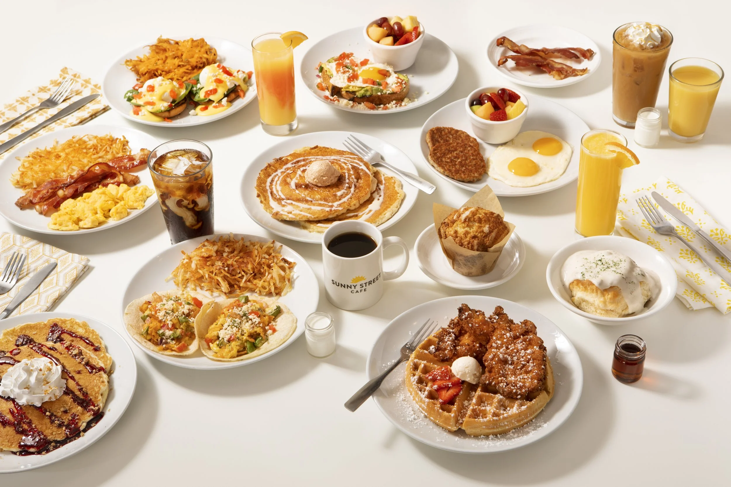An abundant breakfast spread with pancakes, waffles, eggs, bacon, hash browns, toast with toppings, and assorted fruits, along with various beverages including orange juice, coffee, and soft drinks.