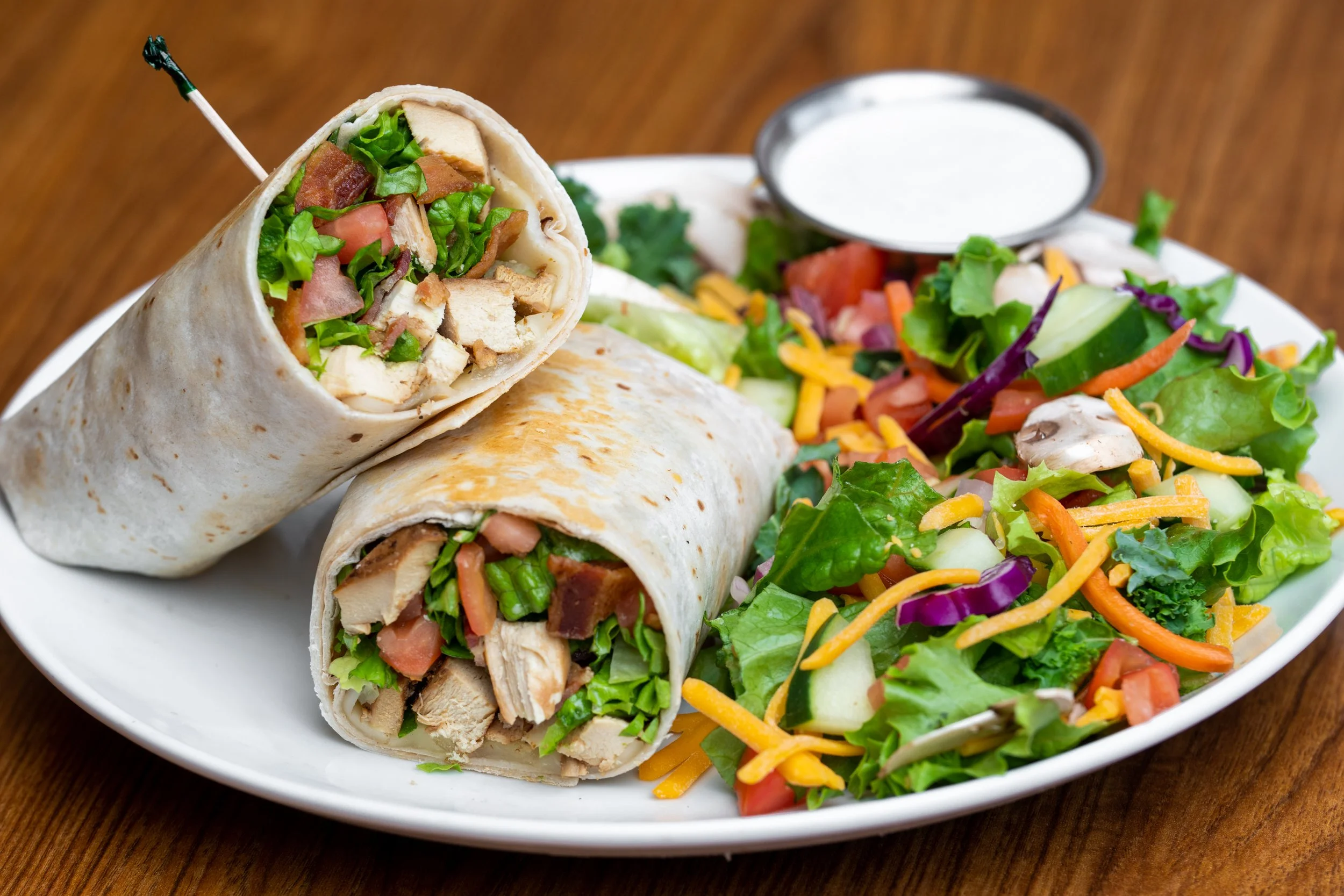 A plate with three chicken wraps filled with lettuce, tomatoes, chicken, and bacon, accompanied by a side salad with shredded cheese, chopped vegetables, and a small cup of white dipping sauce on a wooden table.