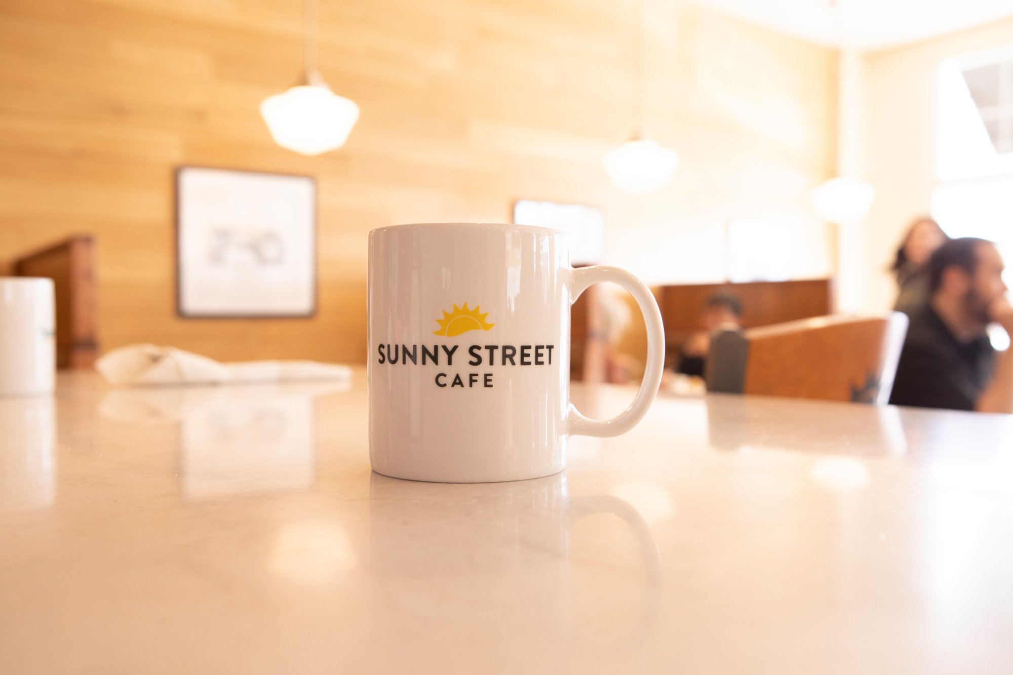 A white coffee mug with the words "Sunny Street Cafe" and a sun graphic on it, placed on a light-colored table inside a cafe with hanging lights and blurred background of people sitting.